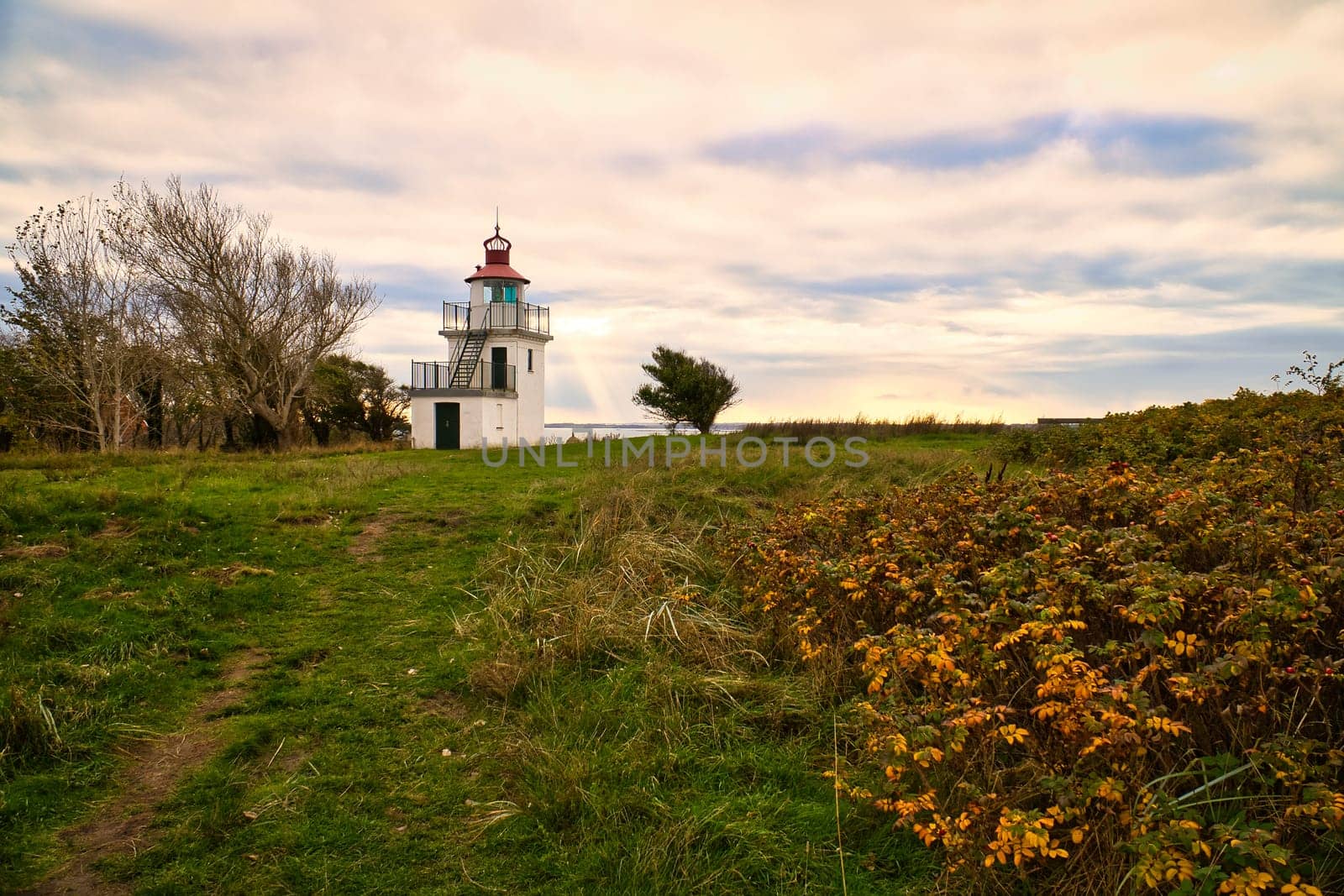 Lighthouse, Spodsbjerg Fyr in Huntsted on the coast of Denmark. Sun rays shining through the clouds. Meadow with trees. Landscape photo from sea