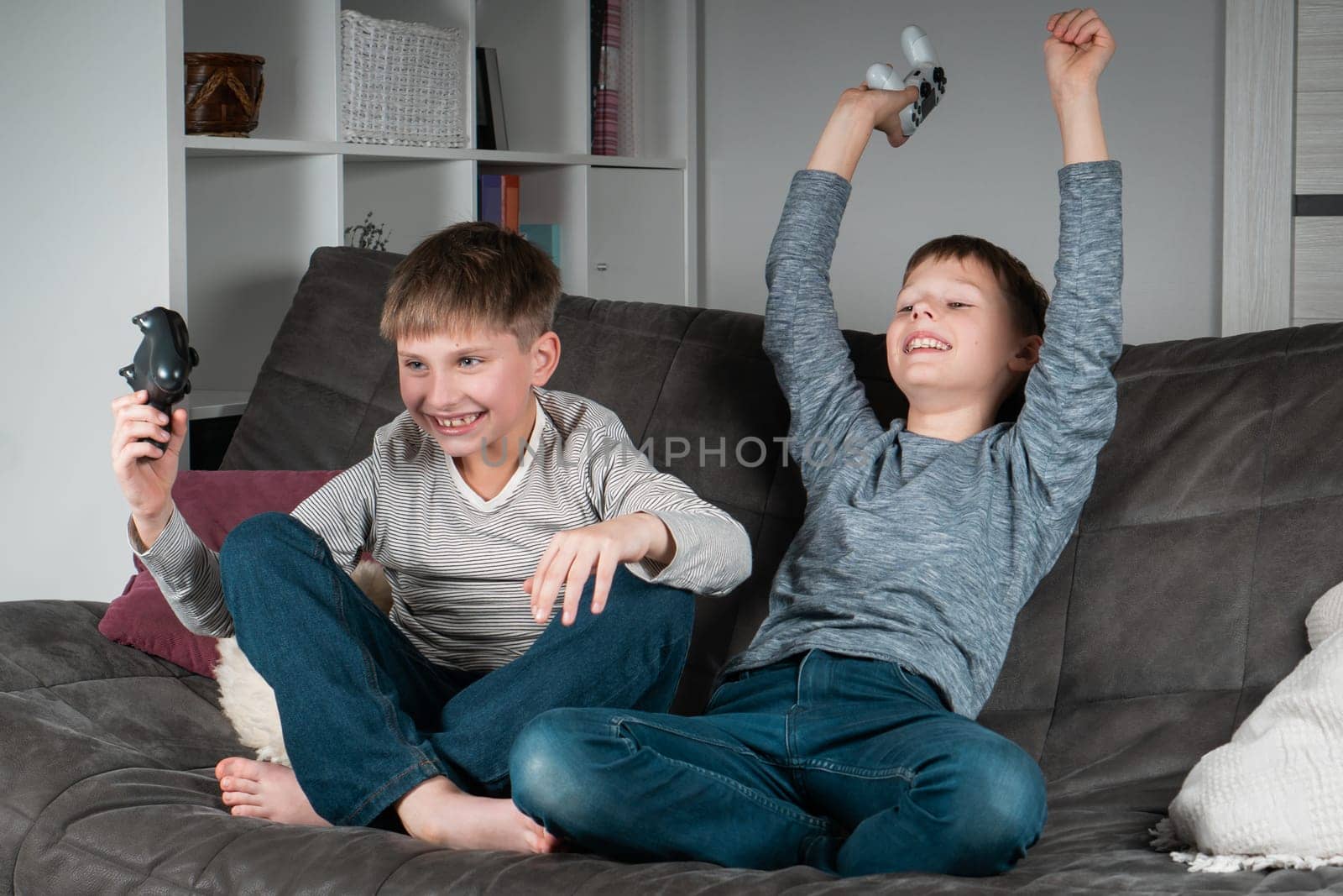 Overjoyed teenagers sit on sofa with game joysticks. Cheerful boys play videogames. Kids having fun together in home room. Modern electronic toys, winning game. Best friends lifestyle.