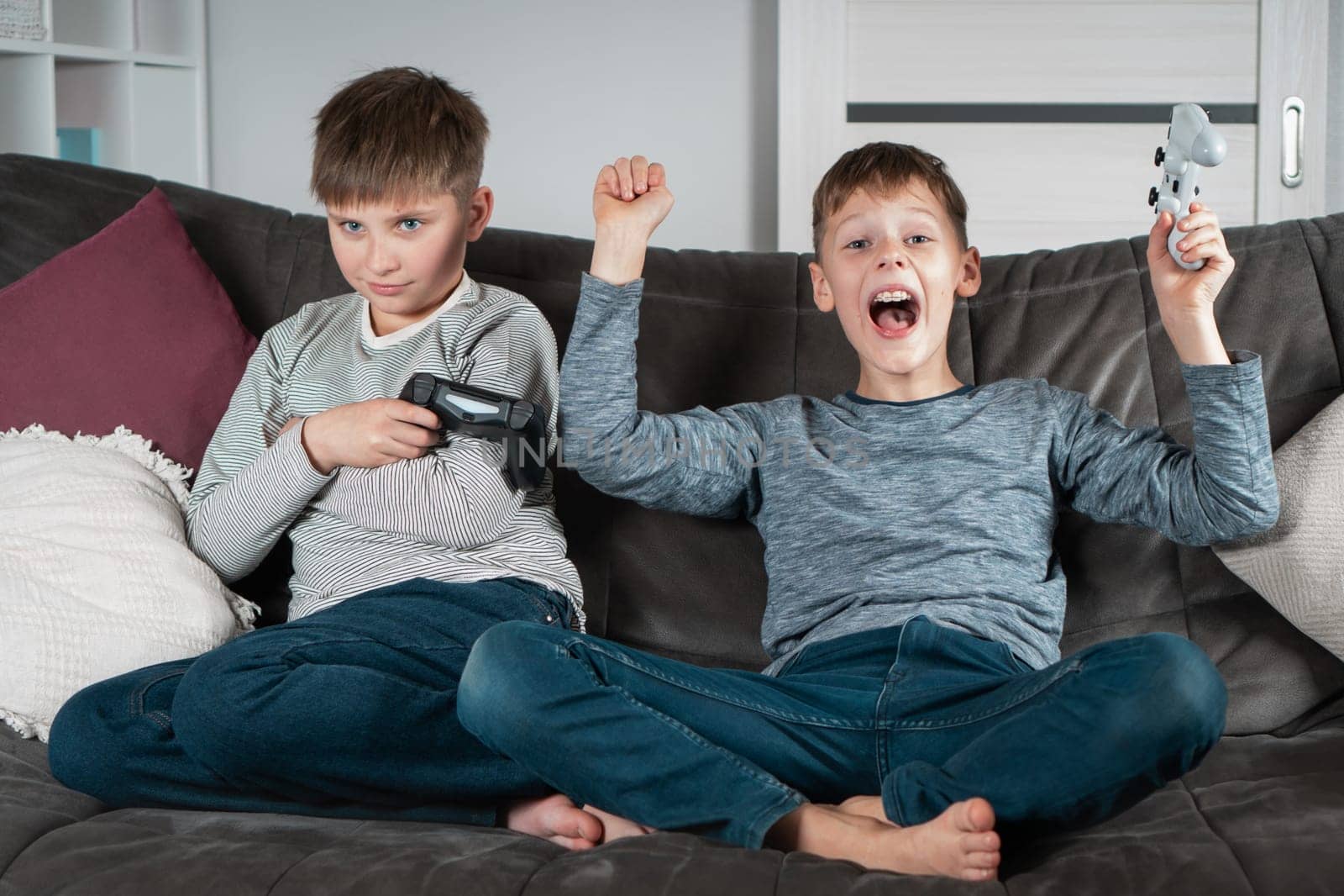Different emotions from winning and losing. School aged boys sit on sofa with joysticks. Children play videogames. Kids having fun together at home. Game competition. Best friends lifestyle.