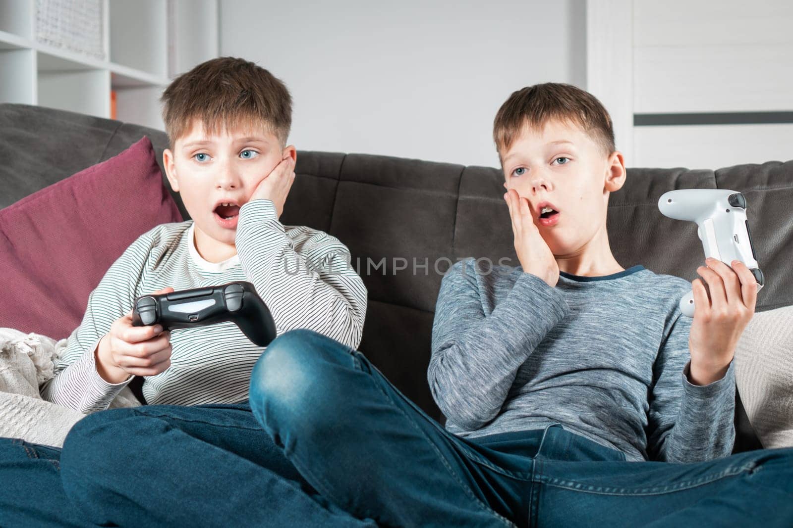 Shocked school aged boys sit on sofa with game joysticks. Children play videogames. Kids relax in home room at daytime together. Disappointment of losing game. Best friends lifestyle.