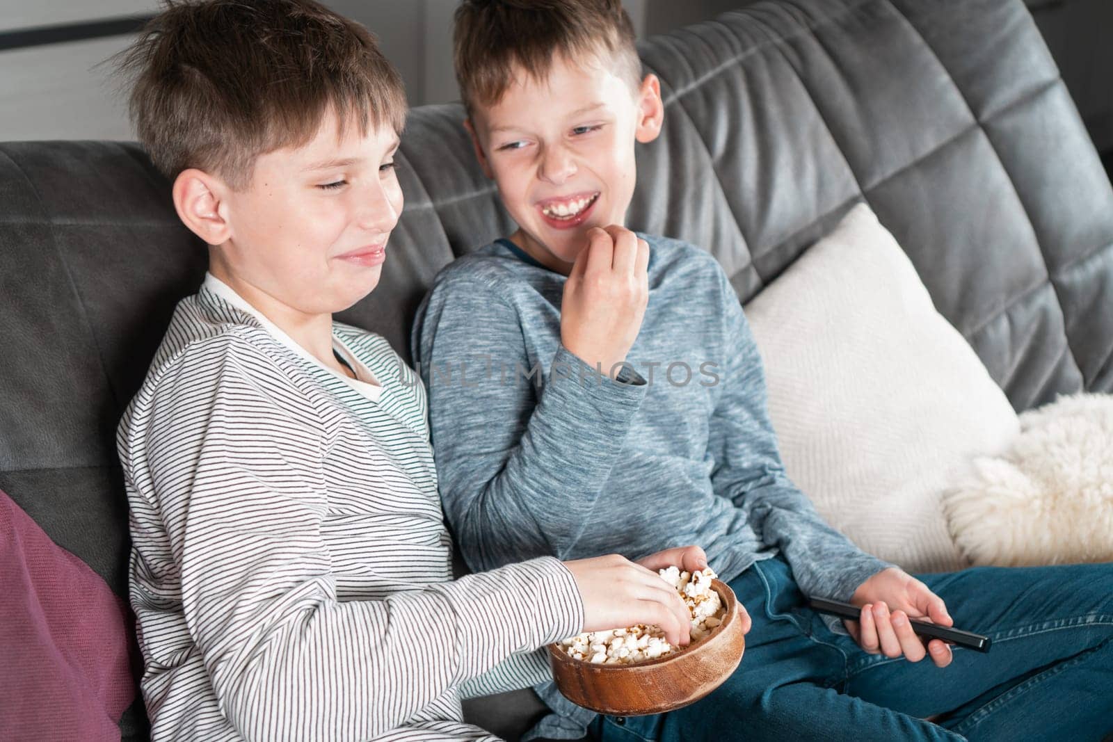 Laughing school aged boys sit on sofa, eat popcorn and watch comic TV show. Overjoyed children relaxing at home. Kids having fun in domestic room at daytime together. Best friends lifestyle.