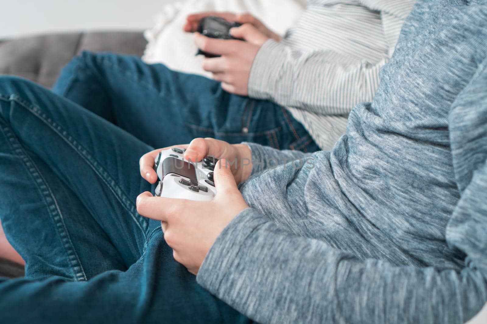 Hands of guys sitting on sofa with game joysticks, closeup side view. Children play videogames. Men in denim and casual longsleeves relaxing at home. Interesting amusement. Best friends lifestyle.
