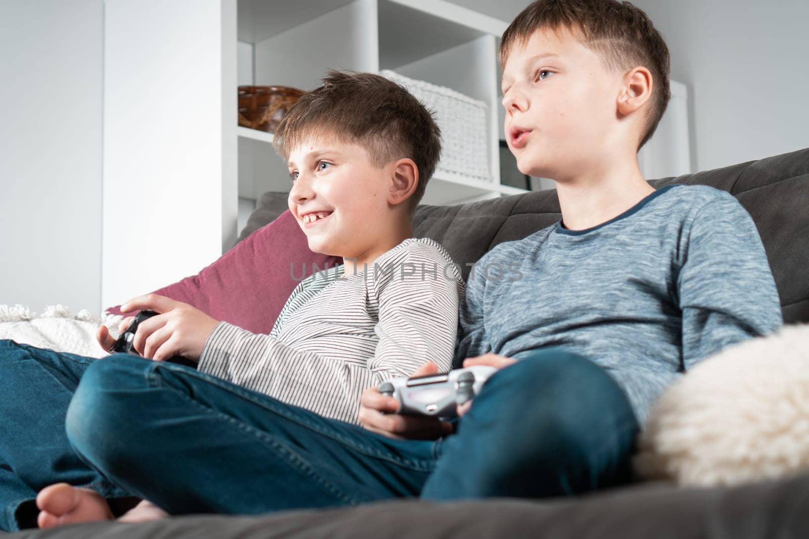 School aged boys sit on sofa with game joysticks, from below view. Children play videogames. Kids having fun in home room at daytime together. Interesting amusement. Best friends lifestyle.