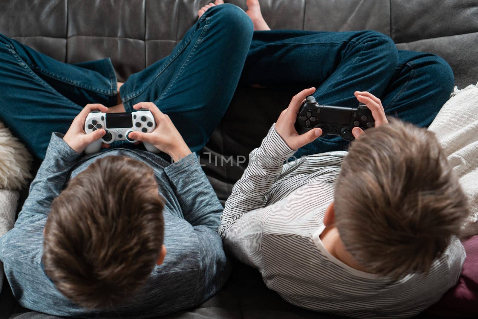 Guys sitting on sofa with game joysticks, from above view of crown and occiput. Children play videogames. Kids in denim and longsleeves relax at home. Interesting amusement. Best friends lifestyle.