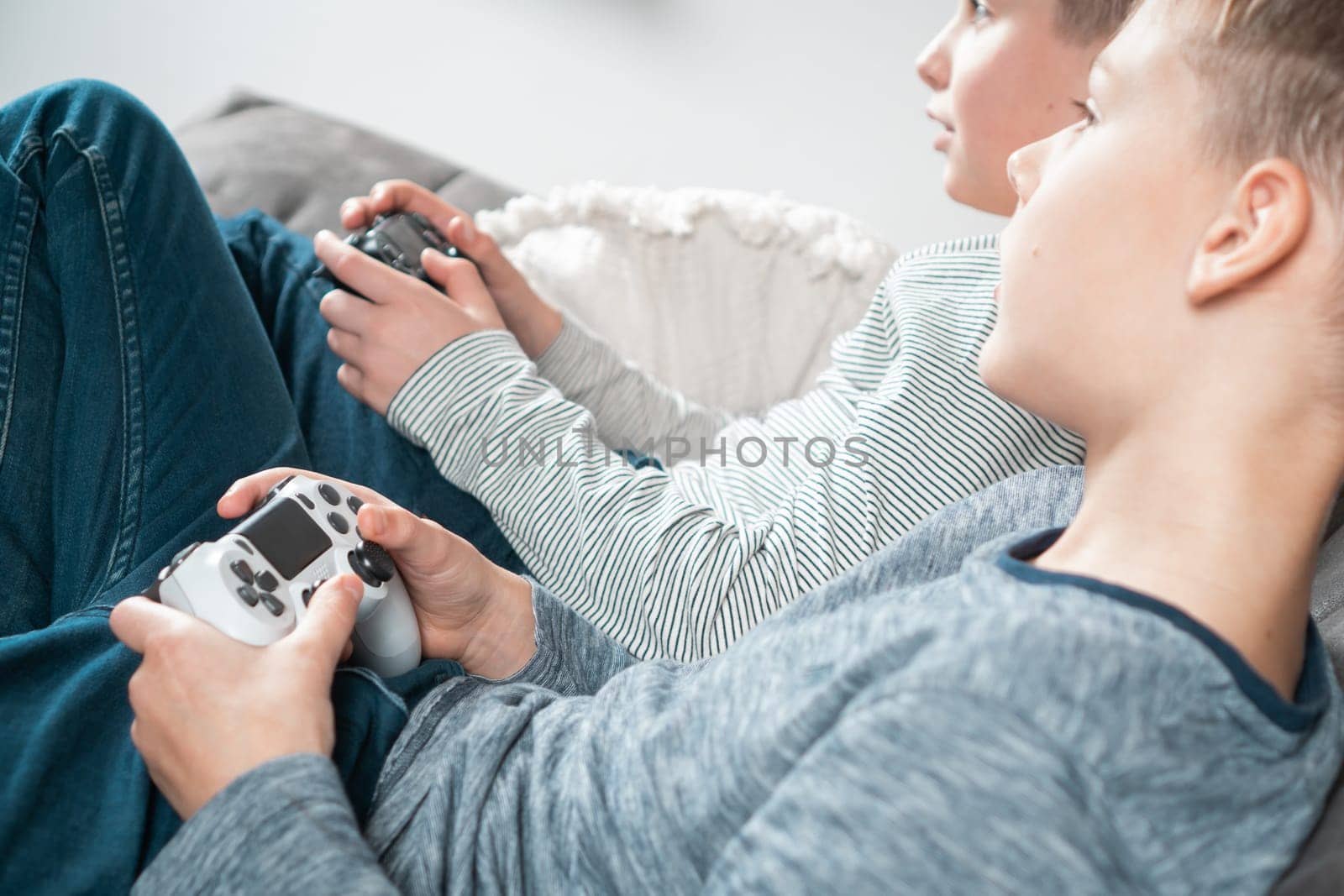 School aged boys sit on sofa with game joysticks, side view from above. Children play videogames. Kids having fun in home room at daytime together. Interesting amusement. Best friends lifestyle.