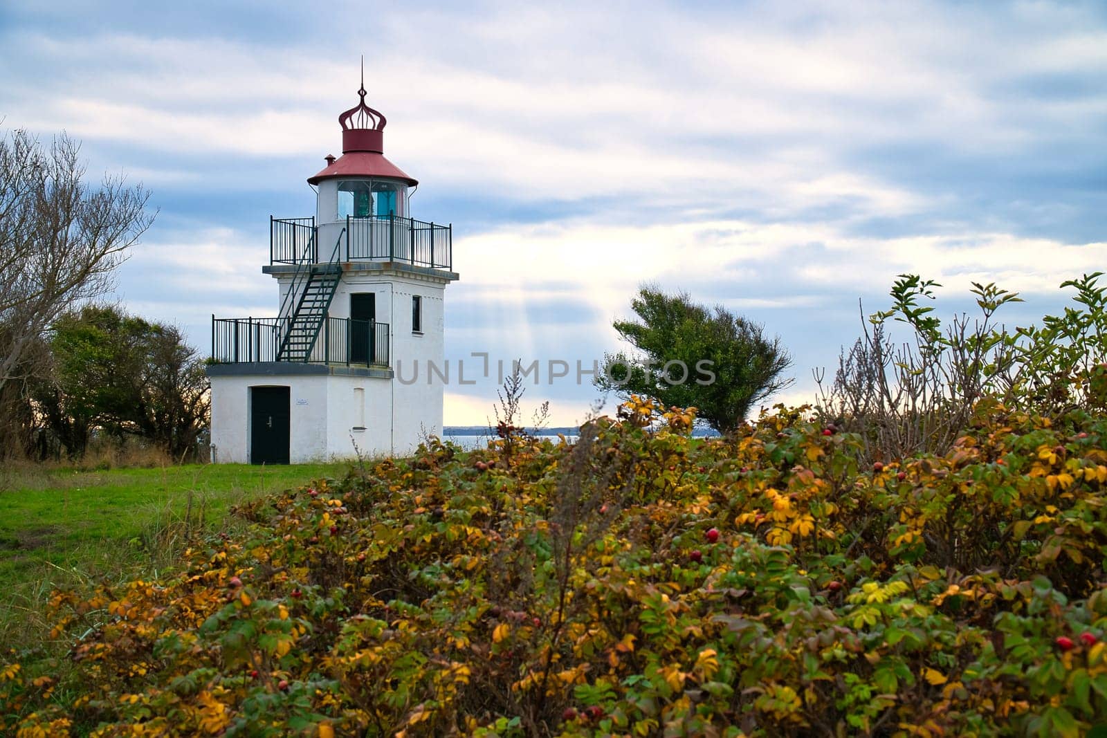 Lighthouse, Spodsbjerg Fyr in Huntsted on the coast of Denmark. Sun rays shining through the clouds. Meadow with trees. Landscape photo from sea