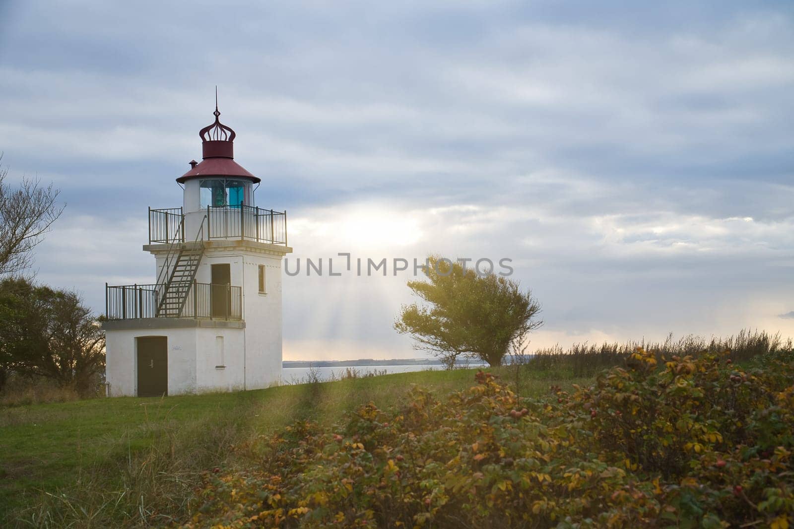 Lighthouse, Spodsbjerg Fyr in Huntsted on the coast of Denmark. Sun rays shining through the clouds. Meadow with trees. Landscape photo from sea