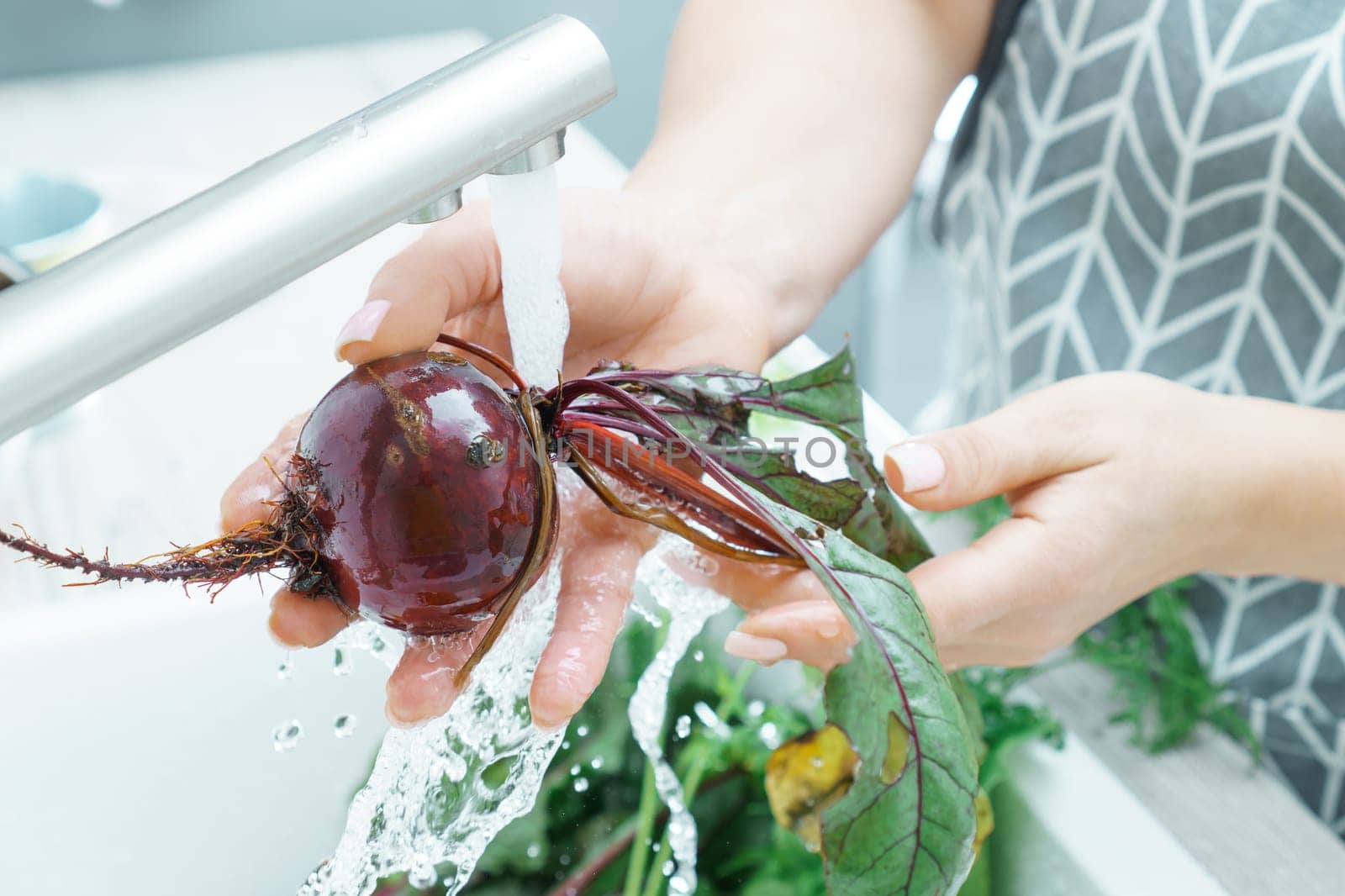 Female hands washing fresh beet under tap water jet in kitchen sink closeup. Dirty raw unpeeled beetroot with green haulm. Cooking, prepare ingredients, food cleaning. Healthy nutrition, vegetables.