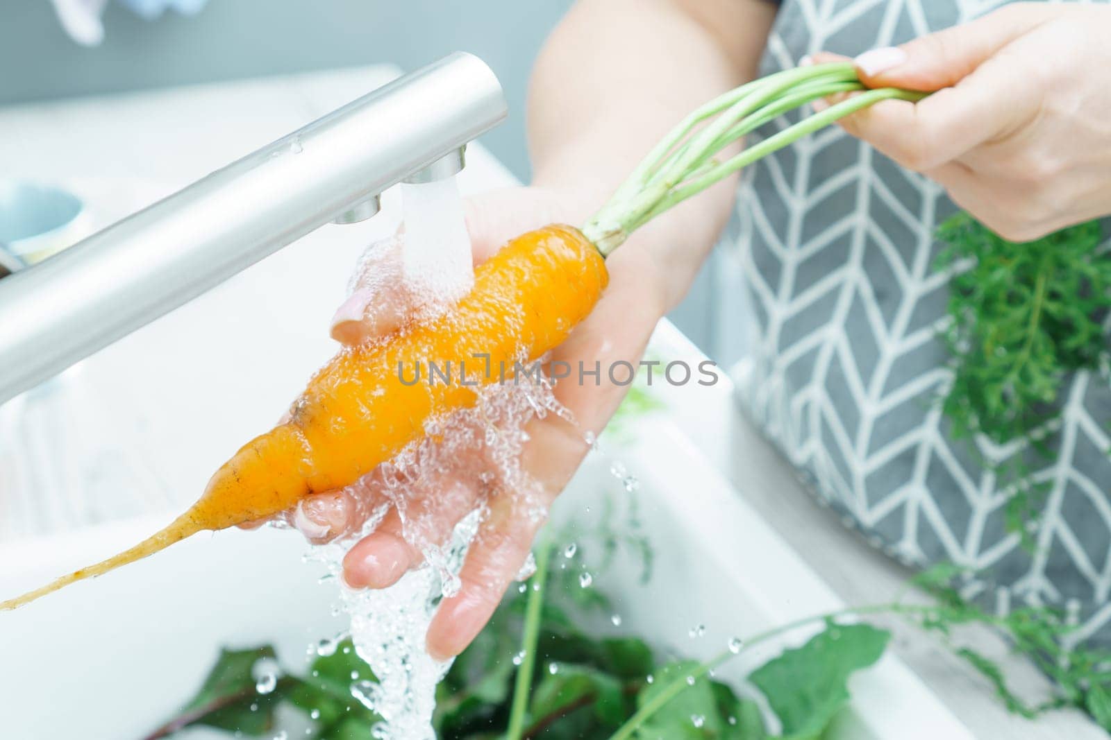 Female hands washing fresh carrot under tap water jet in kitchen sink closeup. Dirty raw orange carrot with green haulm. Cooking, prepare ingredients, food cleaning. Healthy nutrition, vegetables.