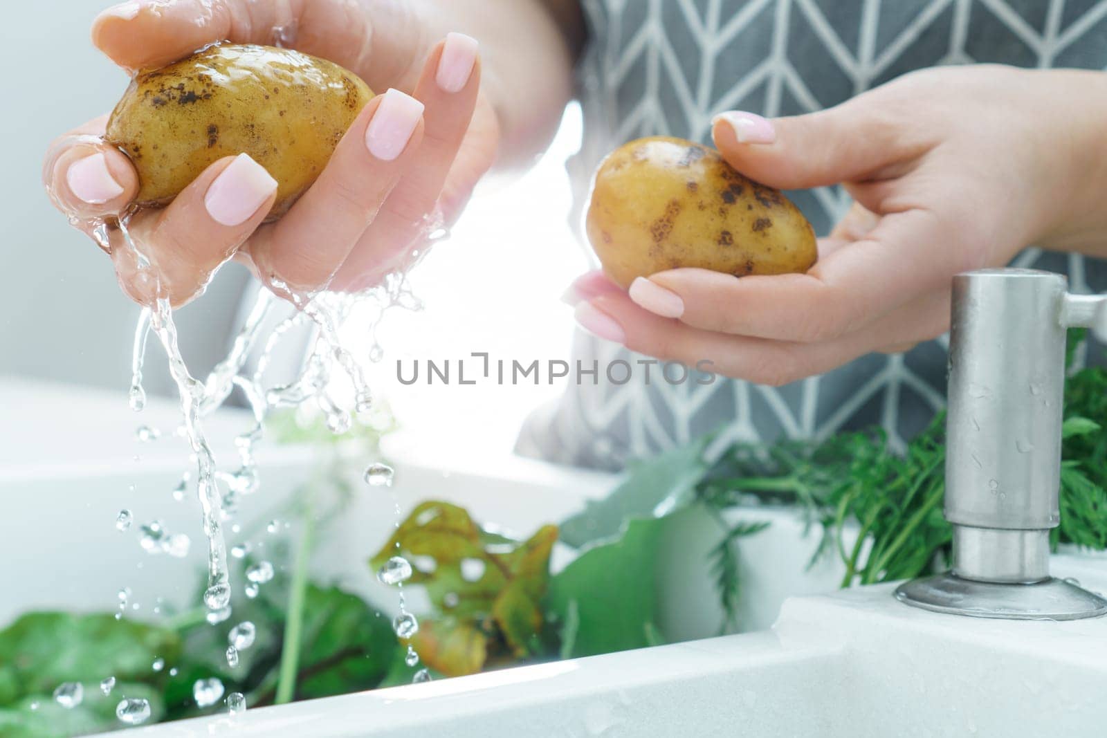 Female manicured hands washing fresh potato under water jet in kitchen sink closeup. Dirty raw vegetables with green haulm. Cooking, prepare ingredients, food cleaning. Healthy nutrition, diet.