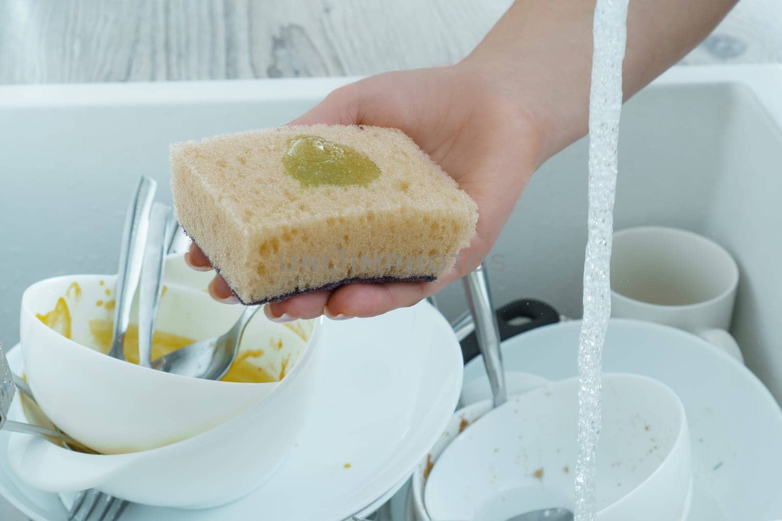 Human hand wash white dinnerware with dish soap on sponge under tap water. Dish washing, tableware after meal, food leftovers. Pile of kitchen utensils in sink closeup. Home cleaning.