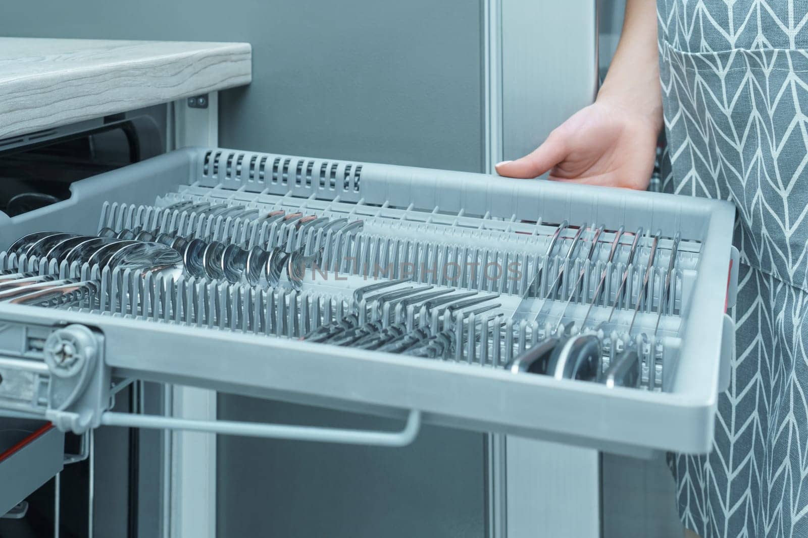 Human hand pull out shelf of dishwasher with clean sparkle cutlery. Dish washing, spoons and forks, household appliance. Modern gray kitchen, silverware cleaning. Convenient kitchen equipment.
