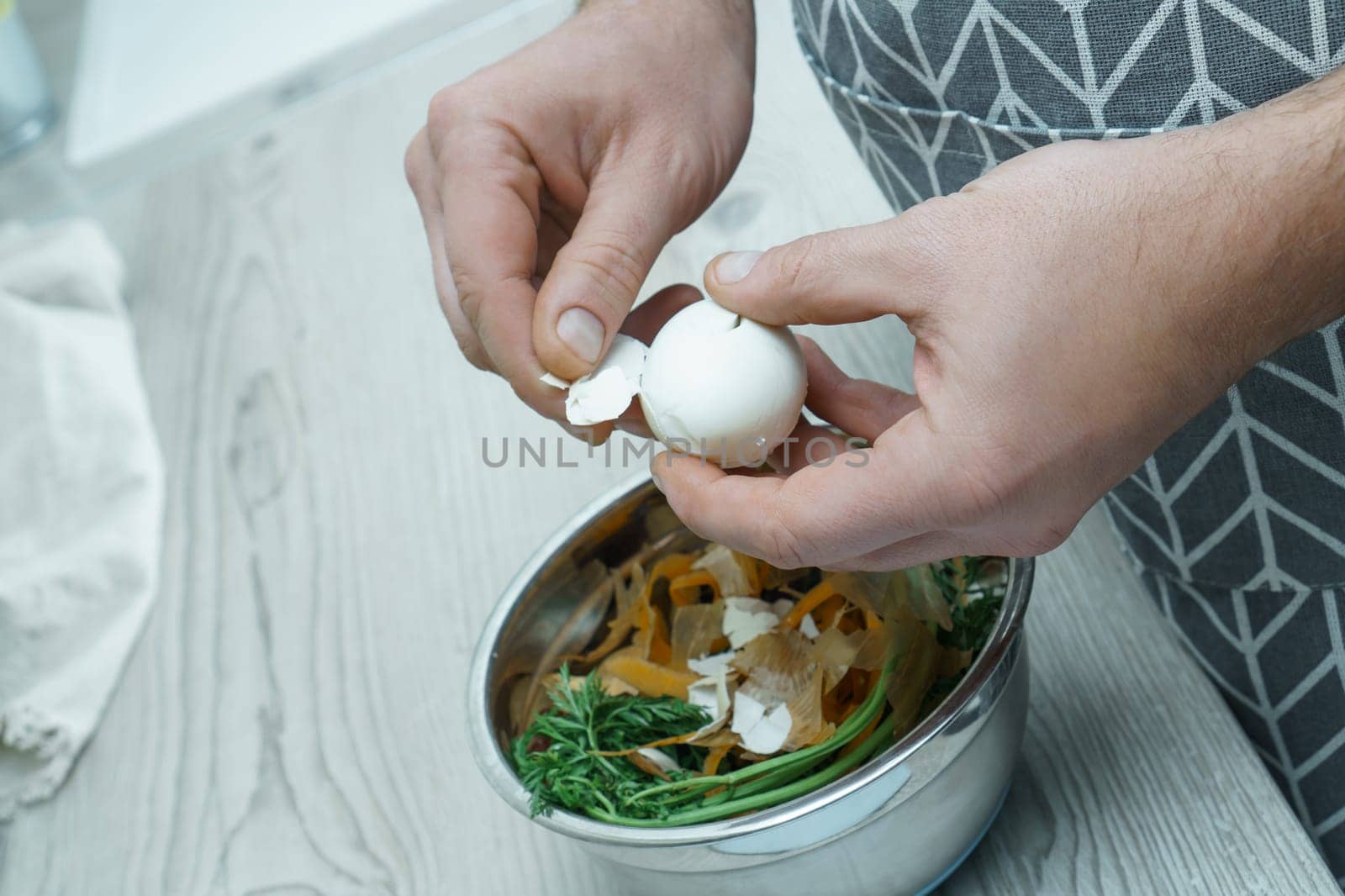 Human hands peeling shell of boiled egg over bowl of vegetable peelings. Person cook on kitchen table, prepare food ingredients. Healthy nutrition, protein product. Separate waste collection.