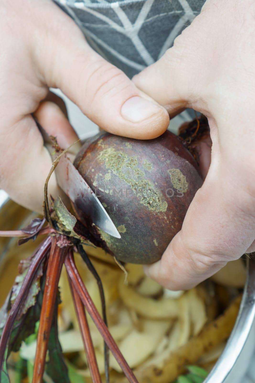 Male hands peeling fresh beetroot and cutting haulm with knife over bowl of vegetable peelings closeup. Prepare ingredients, food scrap. Healthy nutrition, vegetables. Separate waste collection.