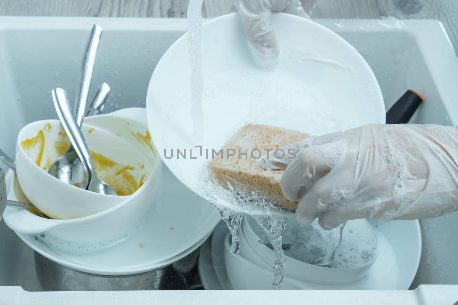 Human hands in protective domestic gloves wash white plate with sponge under tap water. Dish washing, tableware after meal, food leftovers. Pile of kitchen utensils in sink closeup. Home cleaning.