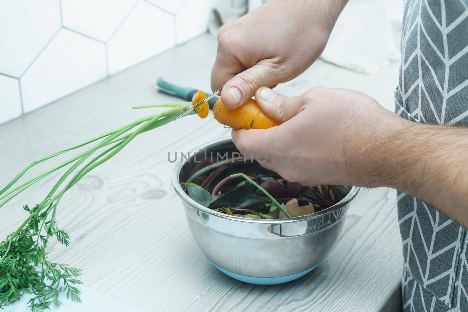 Male hands peeling fresh carrot with knife and cutting haulm over bowl of vegetable peelings. Cook on kitchen table, ingredients, food scrap. Healthy nutrition, vegetables. Separate waste collection.