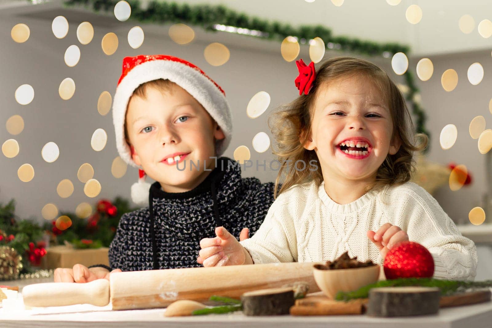 Cute little smiling girl 2-4 with a red bow and boy 7-10 in a Christmas cap, making Christmas gingerbread cookies in the New Year's kitchen.