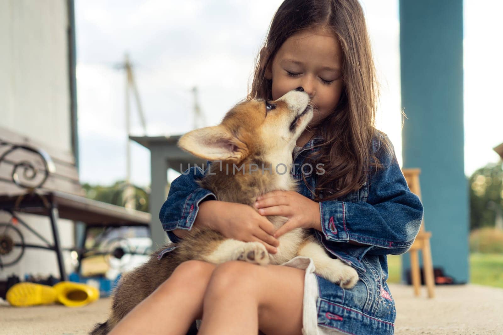 Happy little girl hug and kiss pretty corgi dog sitting outdoors on house yard. Purebred dog, love pets, domestic animal, beautiful small doggy. Child play with brown fluffy cute puppy.