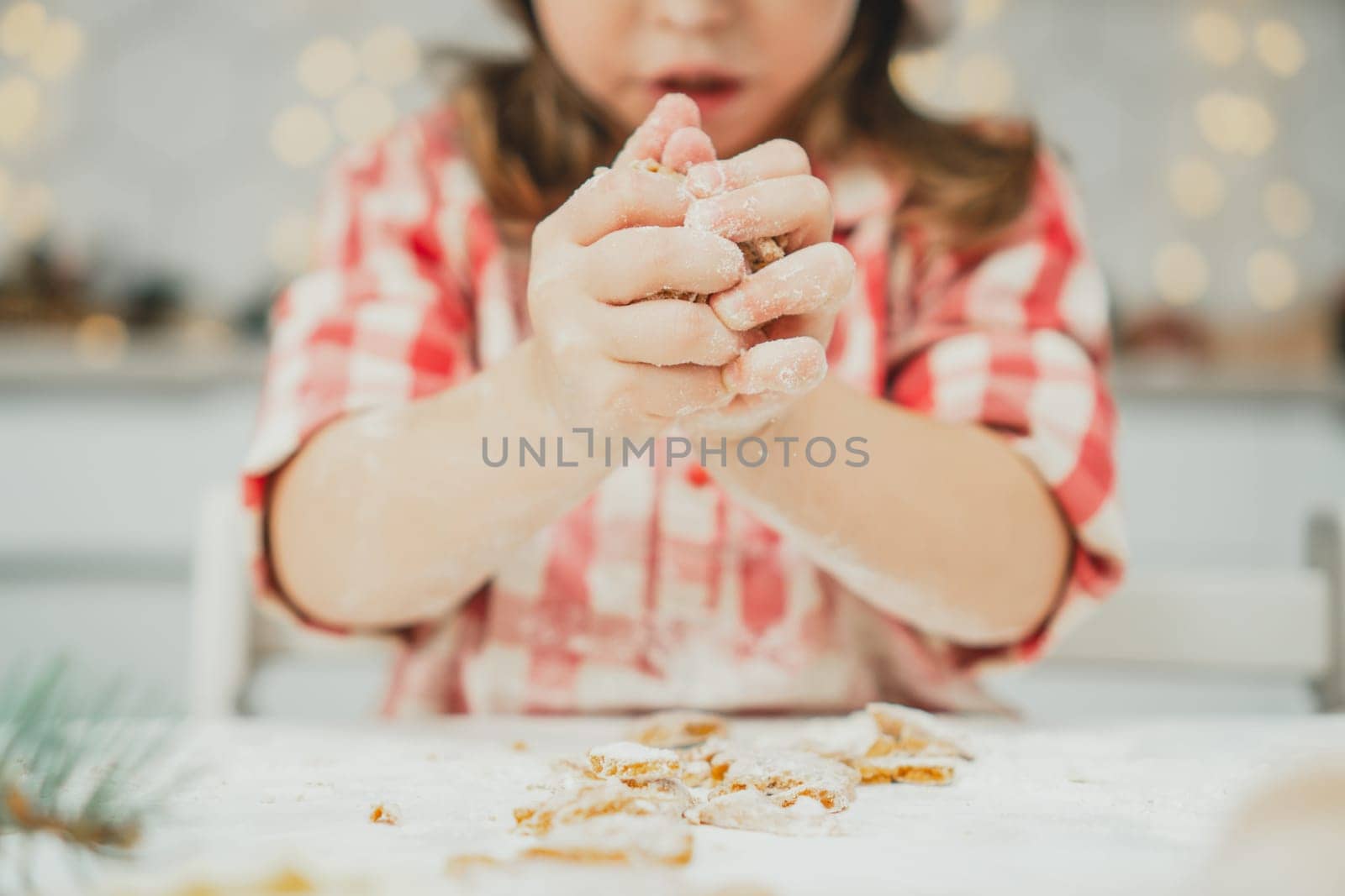 Partially blurred hands of little girl in red checkered shirt prepare dough for gingerbread cookies in white kitchen. Child knead dough on white table strewn with flour