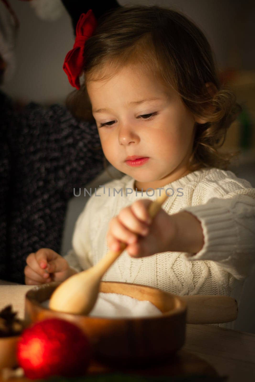Cute little serious girl 2-4 with a red bow prepares Christmas gingerbread cookies in the New Year's kitchen. Vertical.
