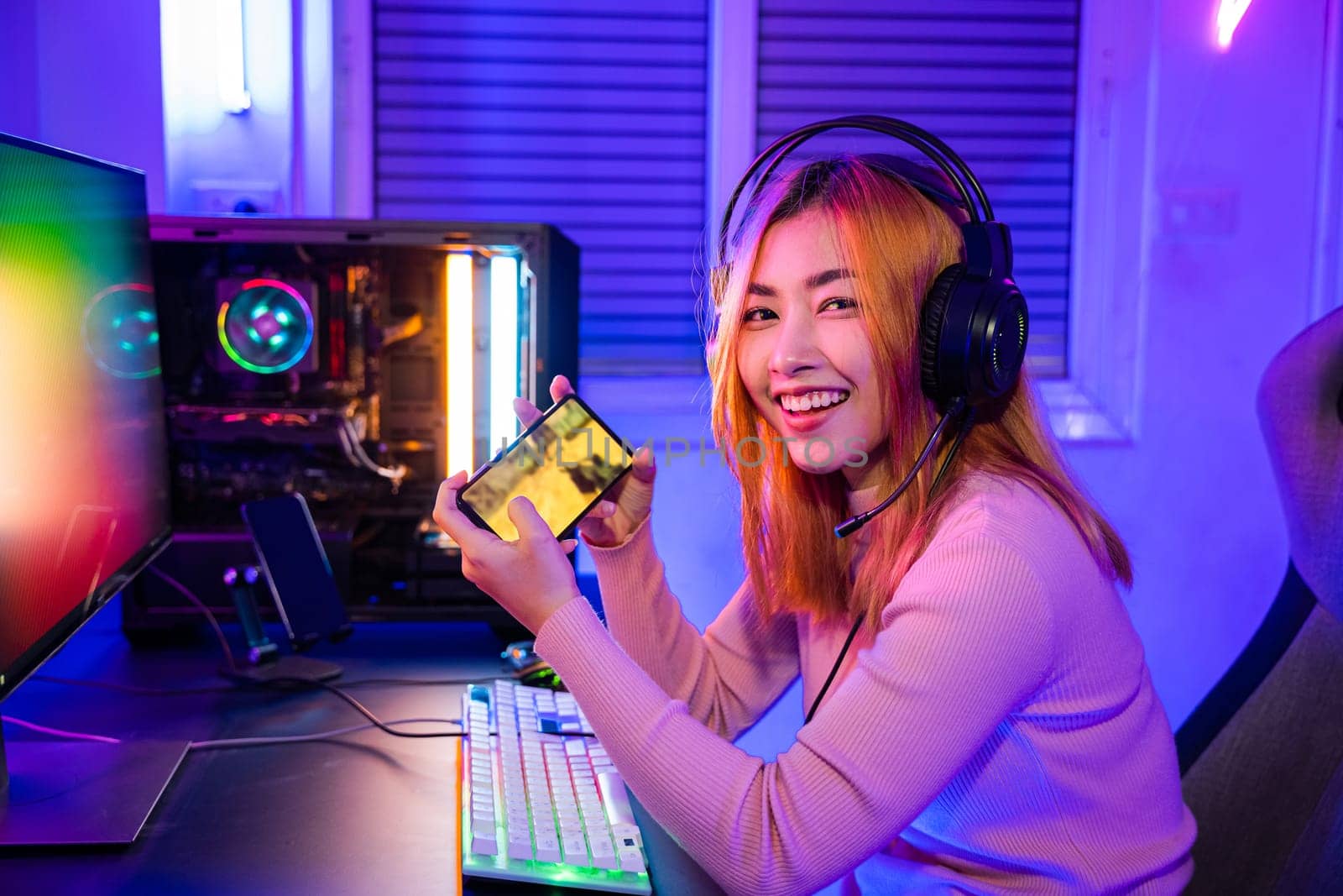 Gamer playing online game application on mobile phone wear gaming headphones looking to camera, Smiling young woman live stream she play video game via smartphone at home neon lights living room