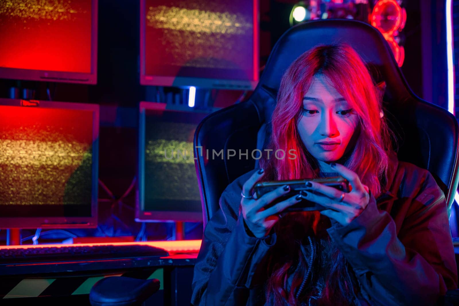 Gamer playing online game application on mobile phone wear gaming headphones, Asian woman live stream she play video game via smartphone at home neon lights living room