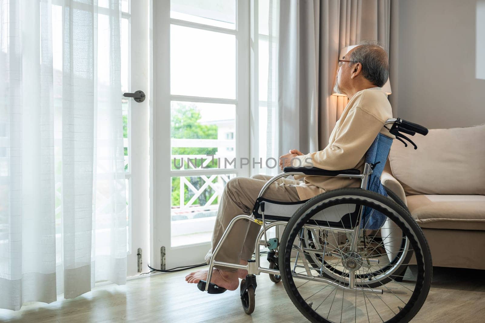 Asian senior man disabled sitting alone in wheelchair looking through window at hospital, lonely elder thoughtful sad old man look outside windows in bedroom at retirement home, feel depressed lonely
