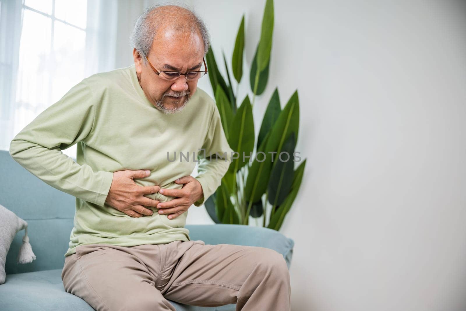 Elderly Asian old man sitting on sofa having stomachache, old age suffering from stomach ache holding his stomach in living room, people health problem, food poisoning