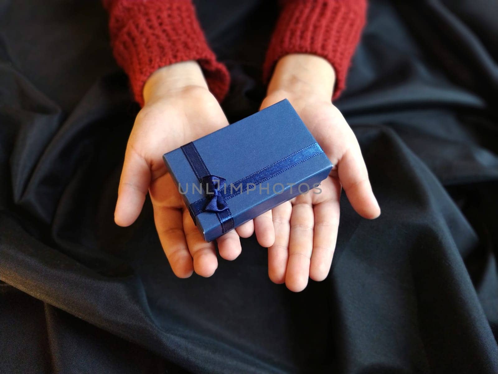 Gift box with a blue bow in children's hands. Black wave-lapped fabric in the background. Gift for New Year, March 8, Birthday, Christmas, Mother's Day or Valentine's Day.