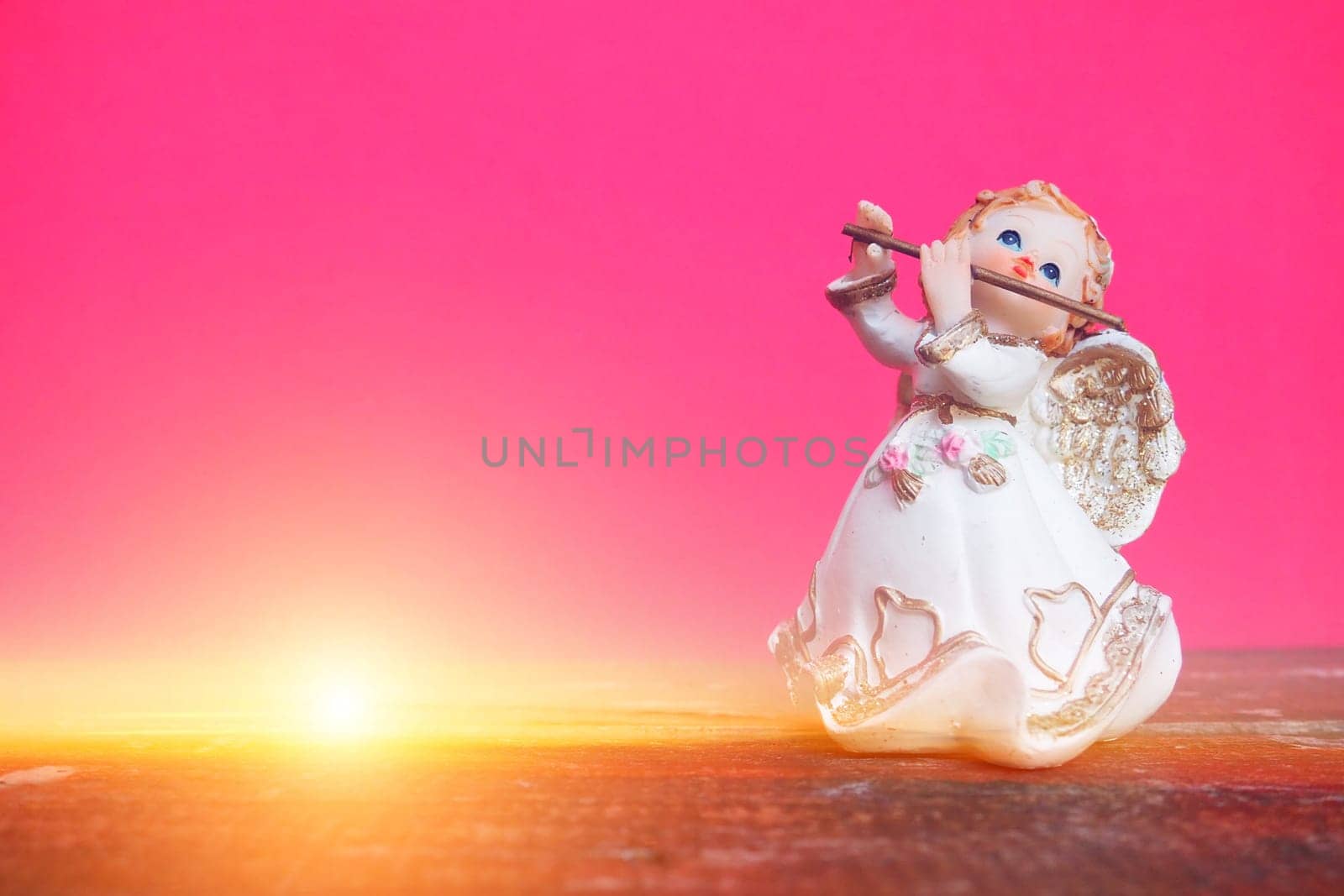 Guardian angel plays the flute. Figurine of a red-haired girl in a white dress with wings and a pipe. New Year or Christmas holiday background. Copy space. Pink wooden background. Sunshine or ray