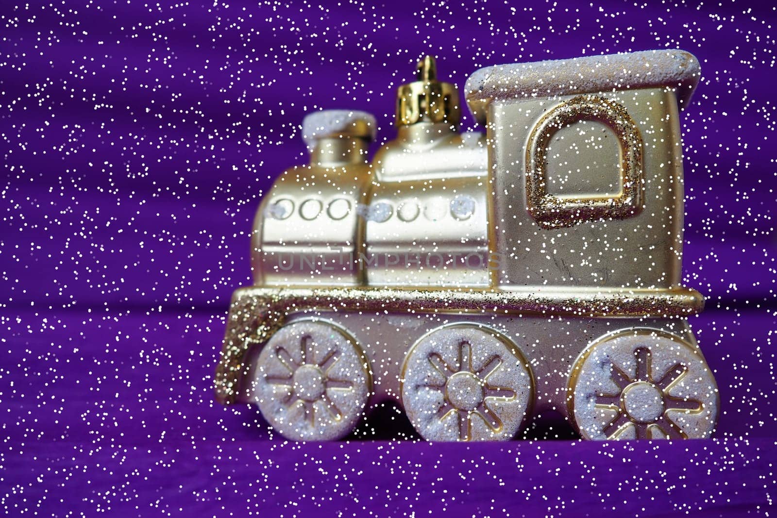 Golden toy train locomotive on a purple and lilac gradient background. New Year or Christmas card. Icicles and snow on the train. Free space for text. Copy space. Festive falling snowflakes bokeh
