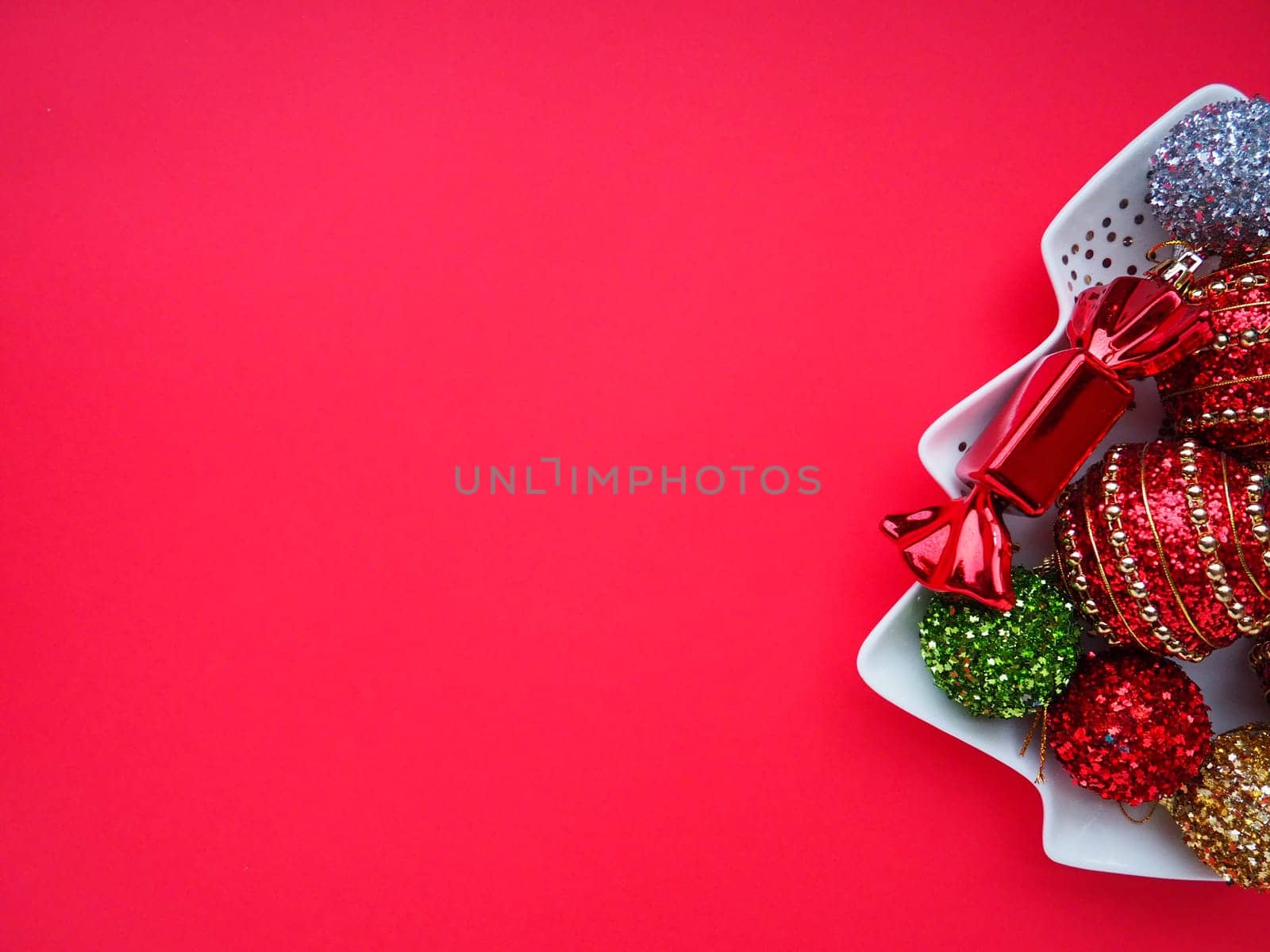 Multicolored Christmas balls and candies on a white plate in the shape of a Christmas tree. Half of the tree is on the right. Copy space Red background. Beautiful striped New Year decor and sparkles.