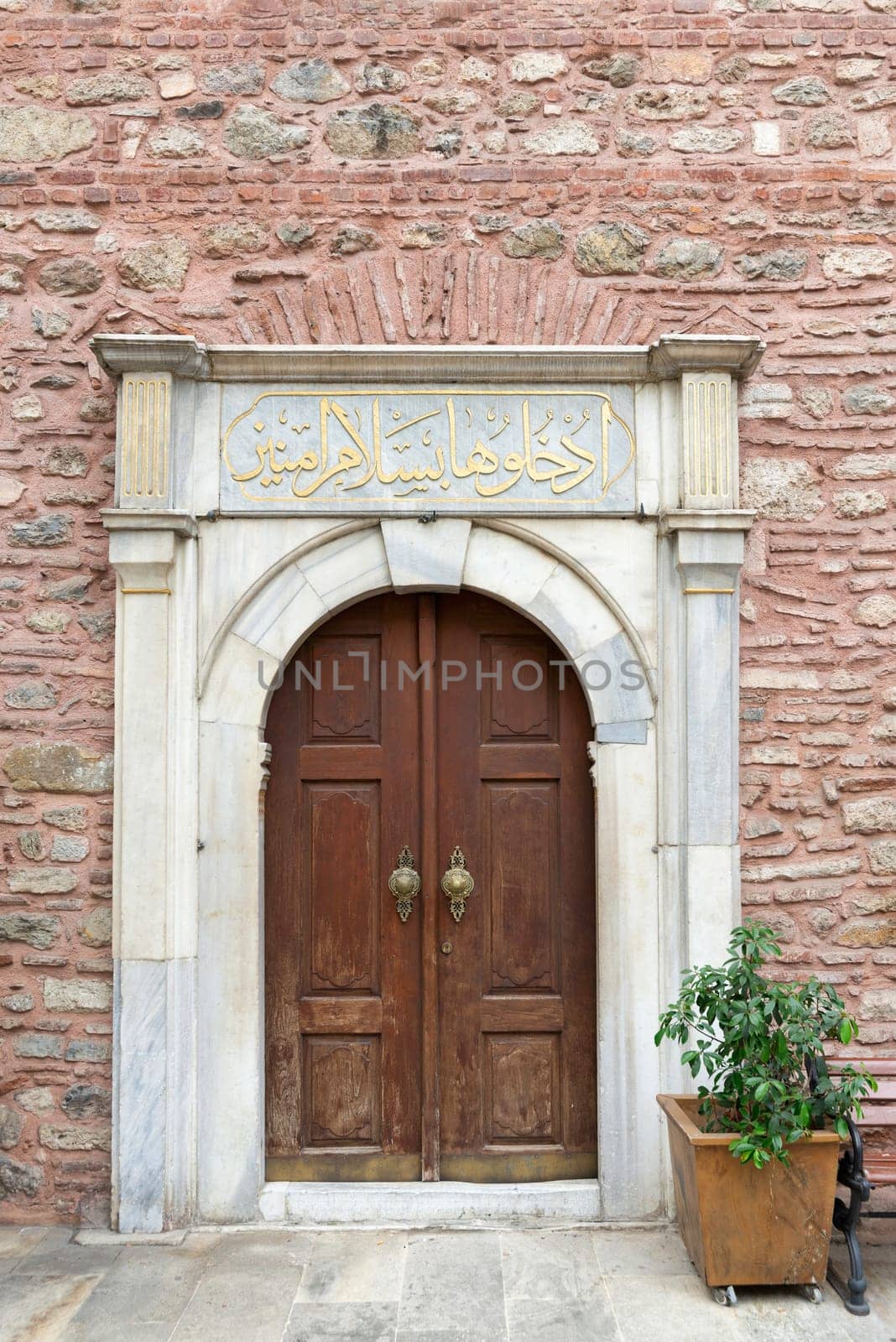 White marble decorated entrance of Arap Mosque, with wooden arched door, in red stone bricks wall, Karakoy, Beyoglu district, Istanbul, Turkey. Text above door says: Enter here in peace, secure