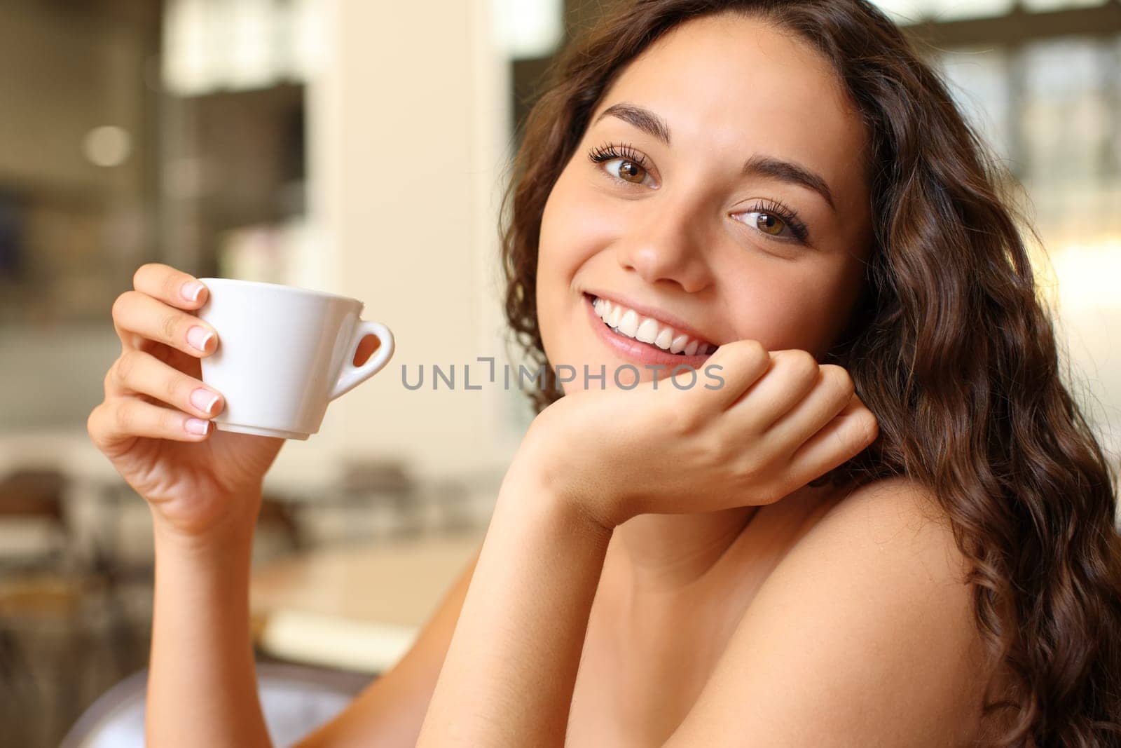Happy woman holding coffee cup looks at you sitting in a bar