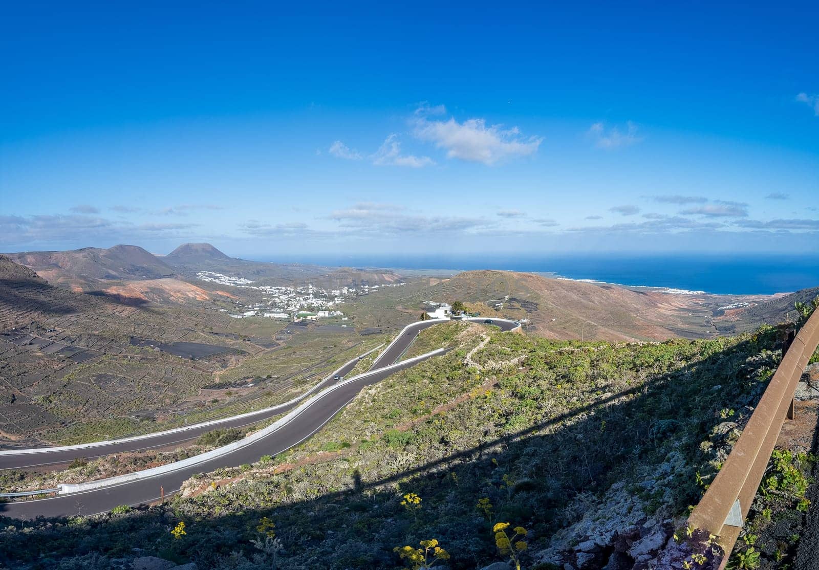 Panoramic view of winding roads in Lanzarote's volcanic landscape, featuring ocean and white villages under a clear blue sky.