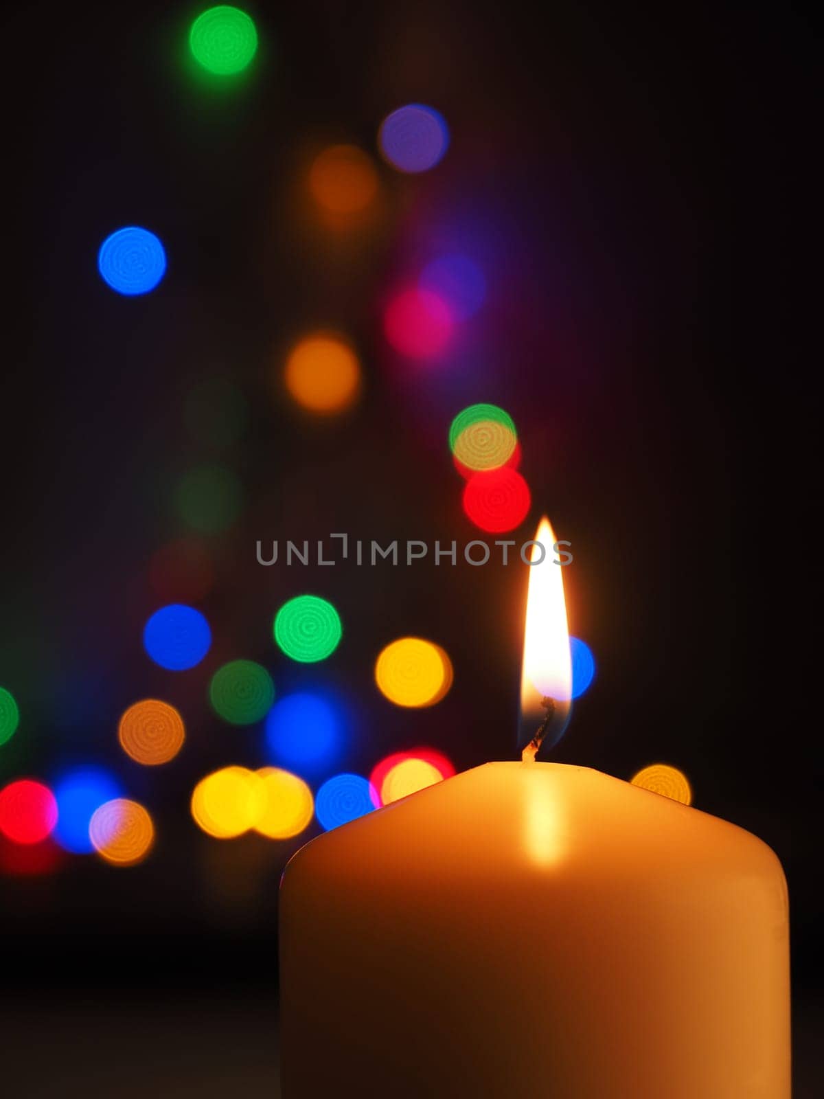 A white candle is burning with colorful Christmas lights bokeh in the background, space for text
