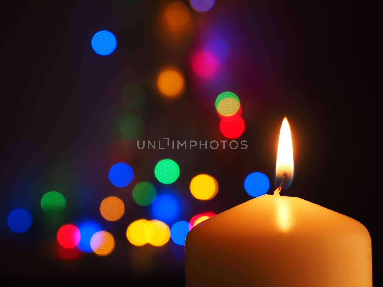 A white candle is burning with colorful Christmas lights bokeh in the background, space for text