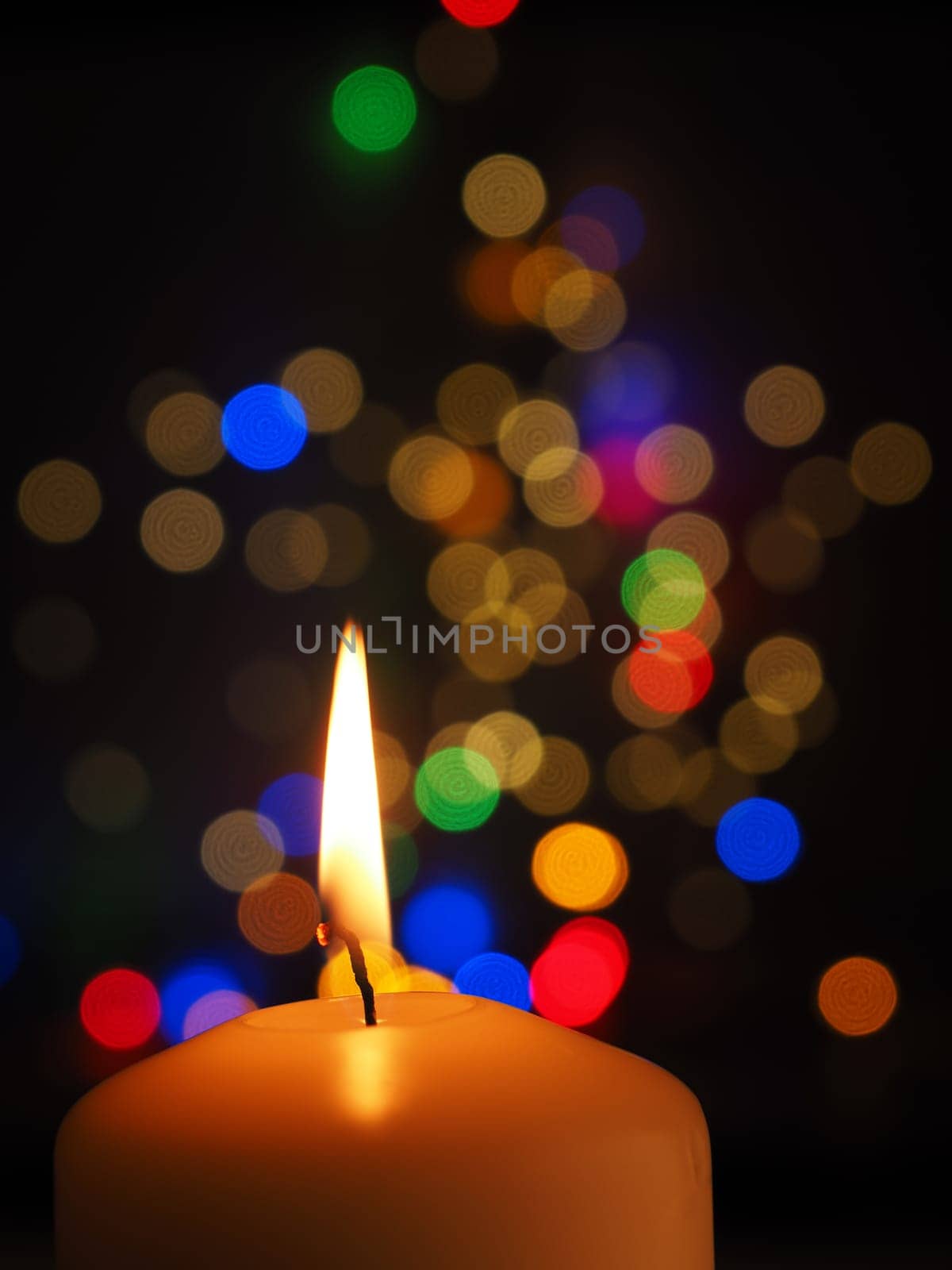 A white candle is burning with colorful Christmas lights bokeh in the background, space for text