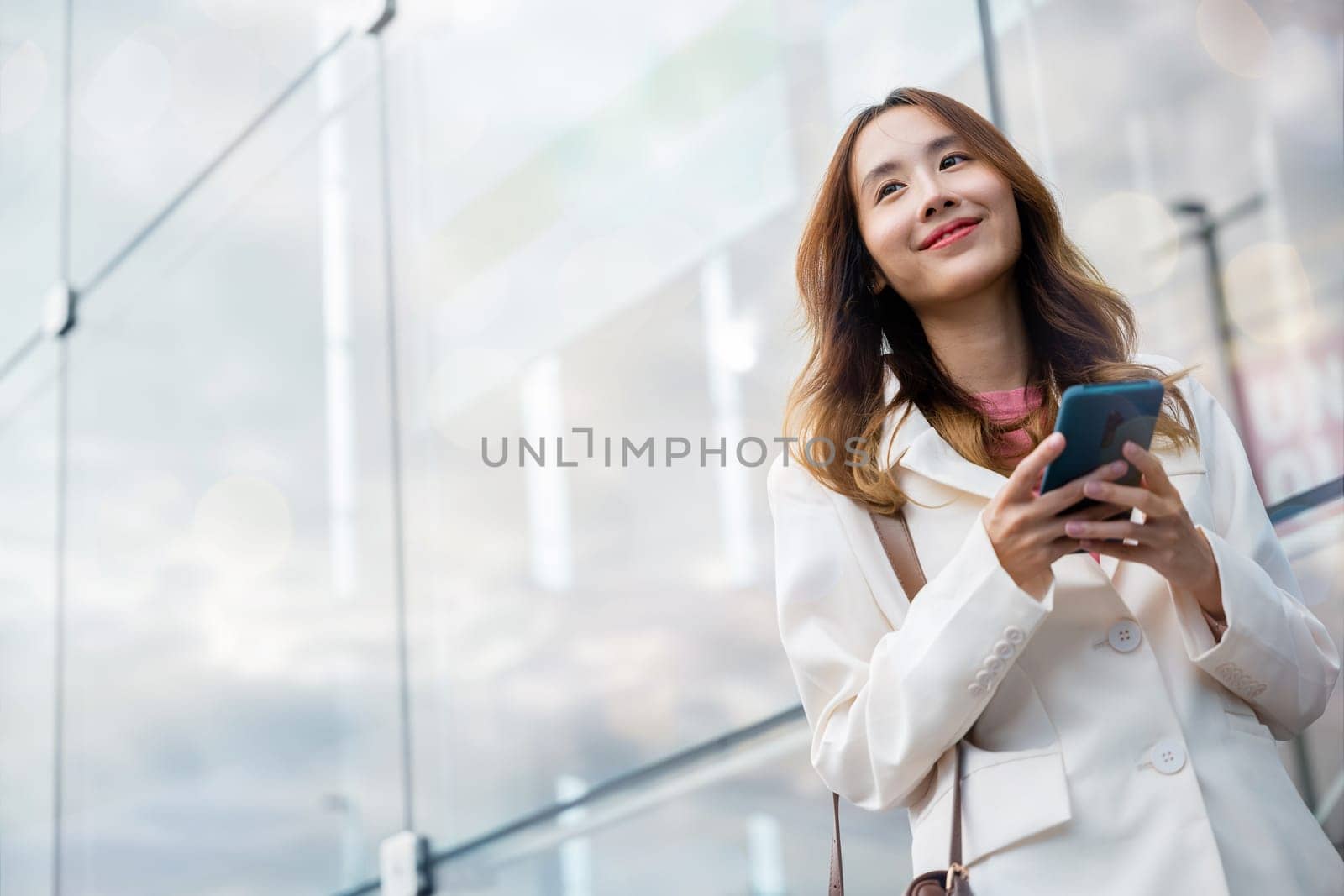 Portrait successful business woman smiling holding smartphone use application chat online in morning, Asian businesswoman working with mobile phone standing against street front building near office