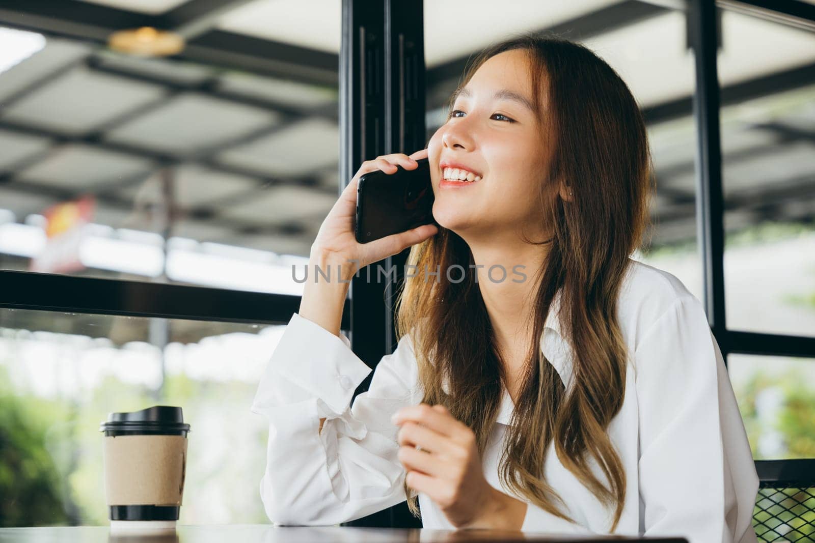Beautiful young businesswoman freelance lunch outside holding smartphone and coffee paper cup at coffee shop, Happy female lifestyle sitting in cafe near window she using mobile phone