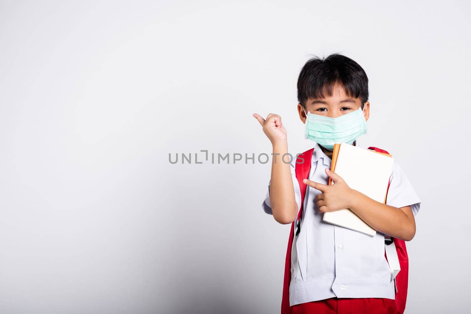 Asian student kid boy wear student thai uniform and protect face mask ready to go to school pointing finger to space in studio shot isolated on white background, preschool, new normal back to school