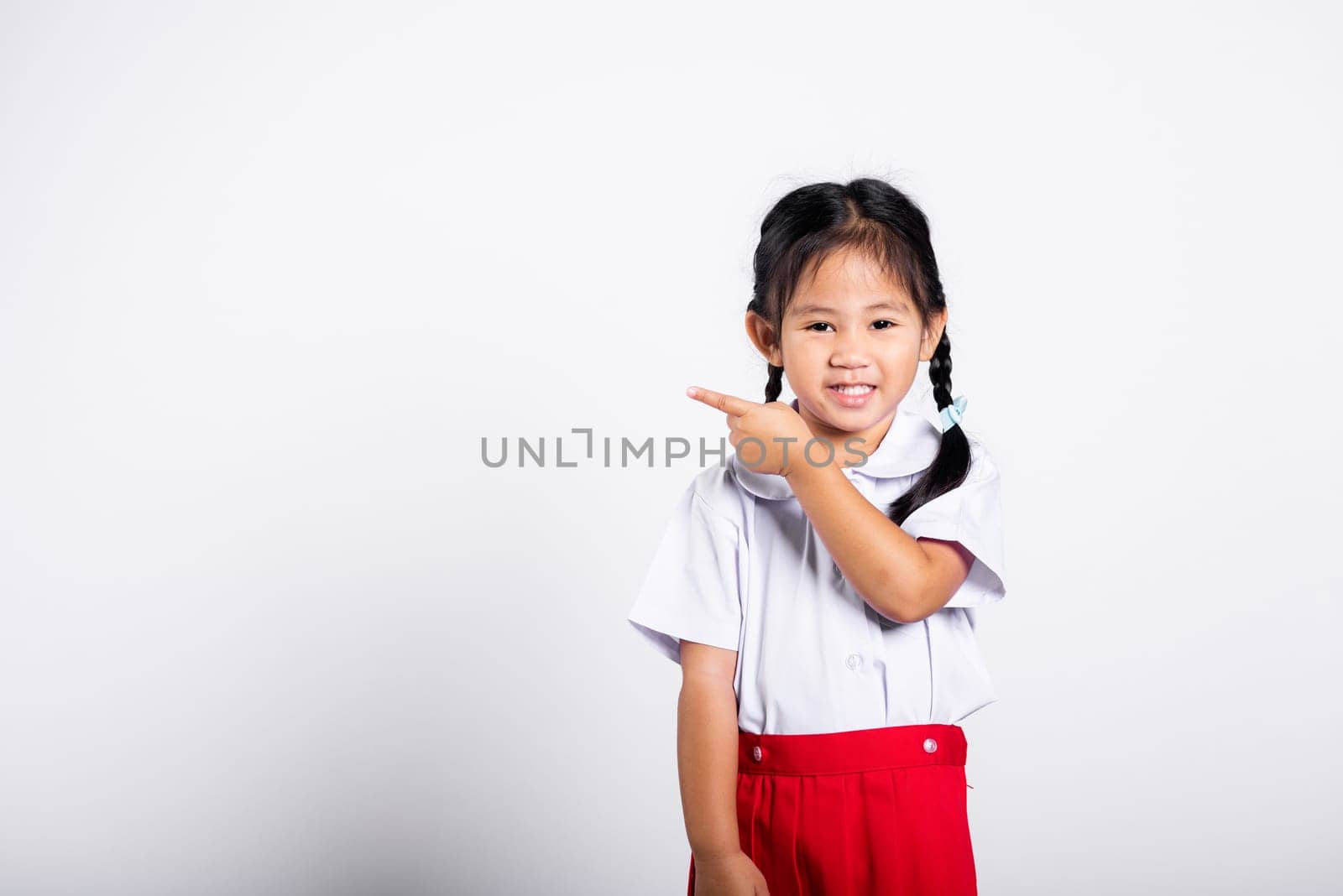 Asian toddler smiling happy wear student thai uniform red skirt keeps pointing finger at copy space in studio shot isolated on white background, Portrait little children girl preschool, Back to school