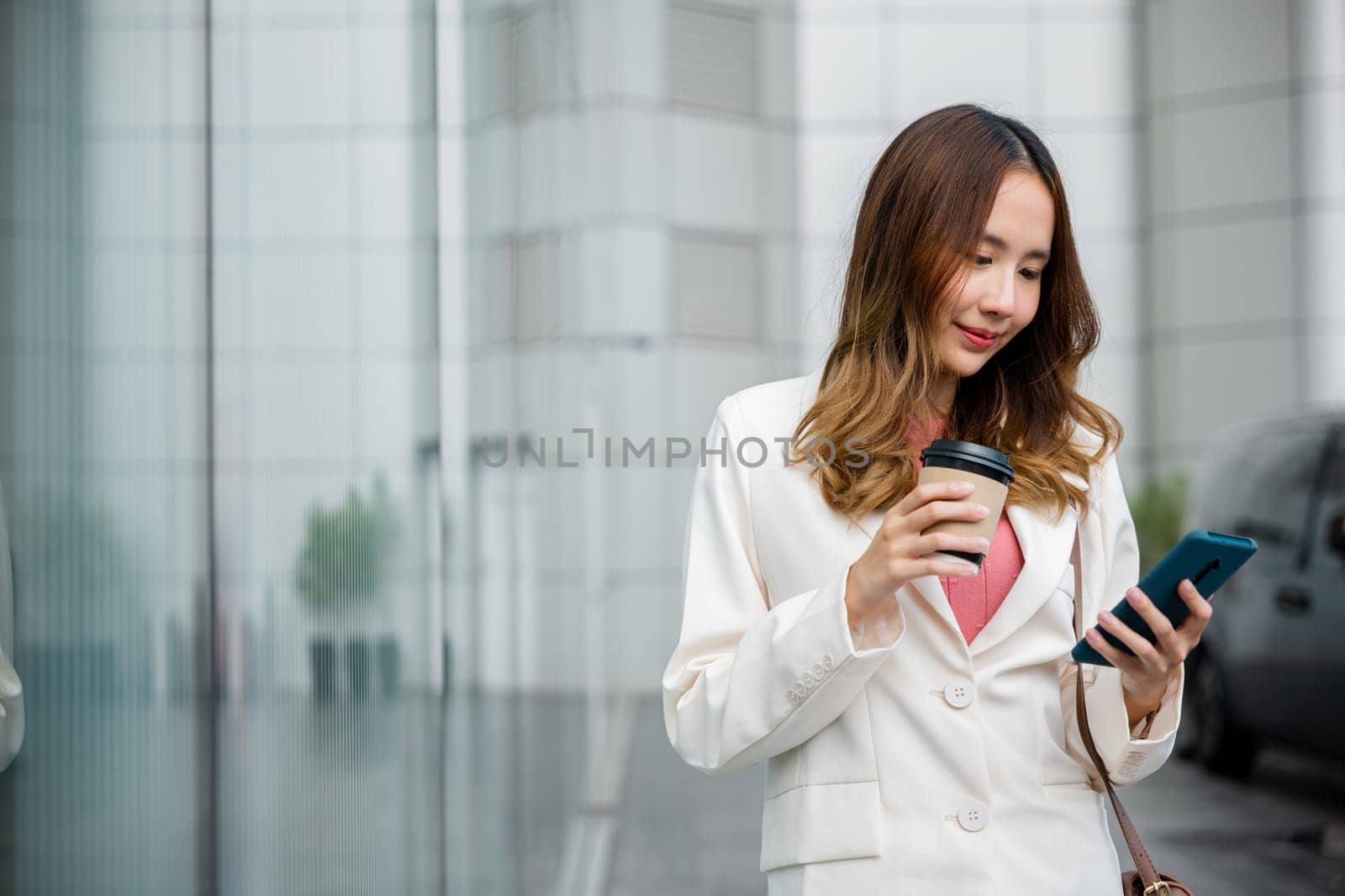 Asian businesswoman holding coffee cup takeaway look smartphone going to work walk on city street near office in morning, Portrait business woman hold mobile phone and paper cup of hot drink outdoor