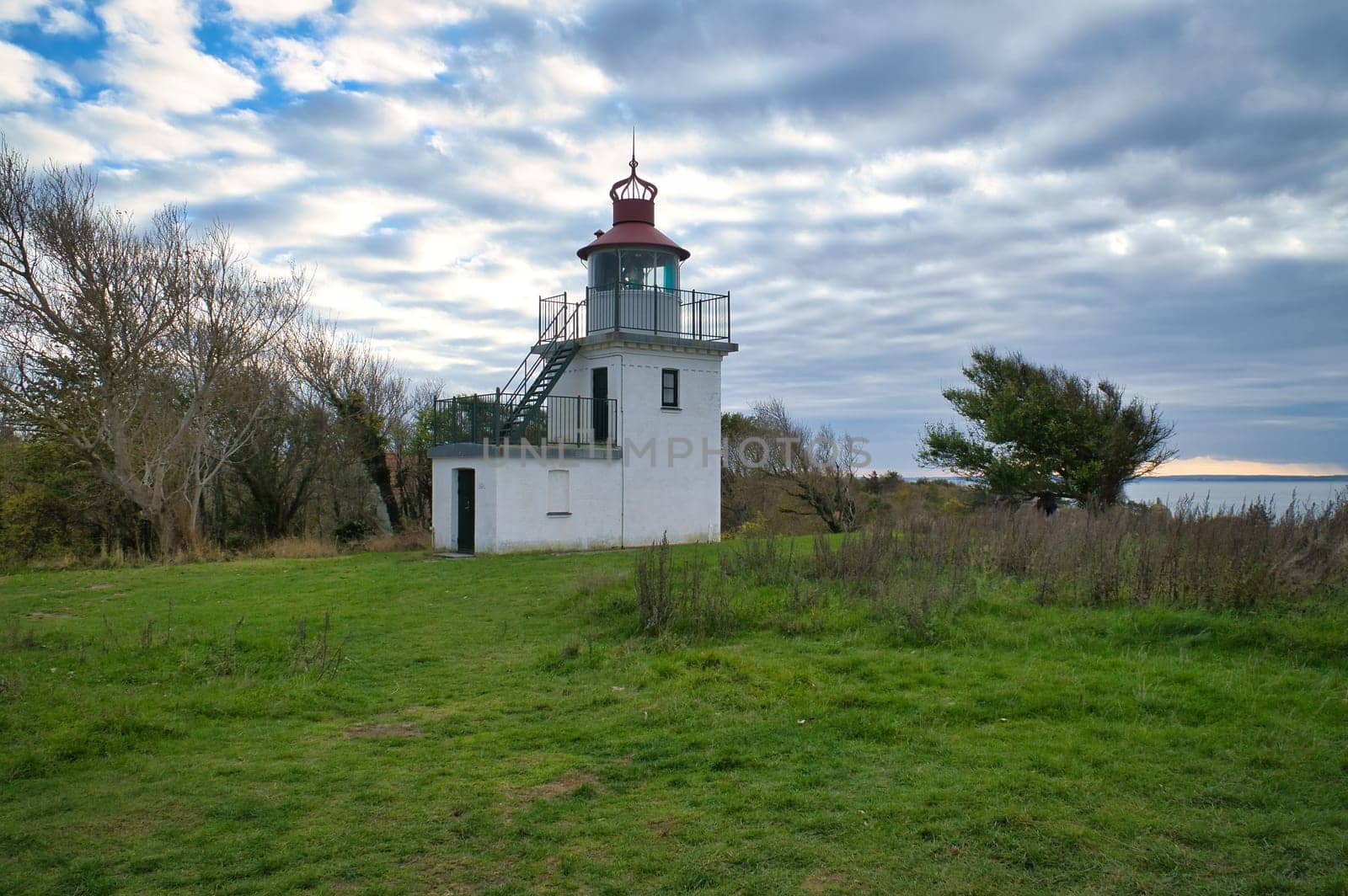 Lighthouse, Spodsbjerg Fyr in Huntsted on the coast of Denmark. Sun rays shining through the clouds. Meadow with trees. Landscape photo from sea