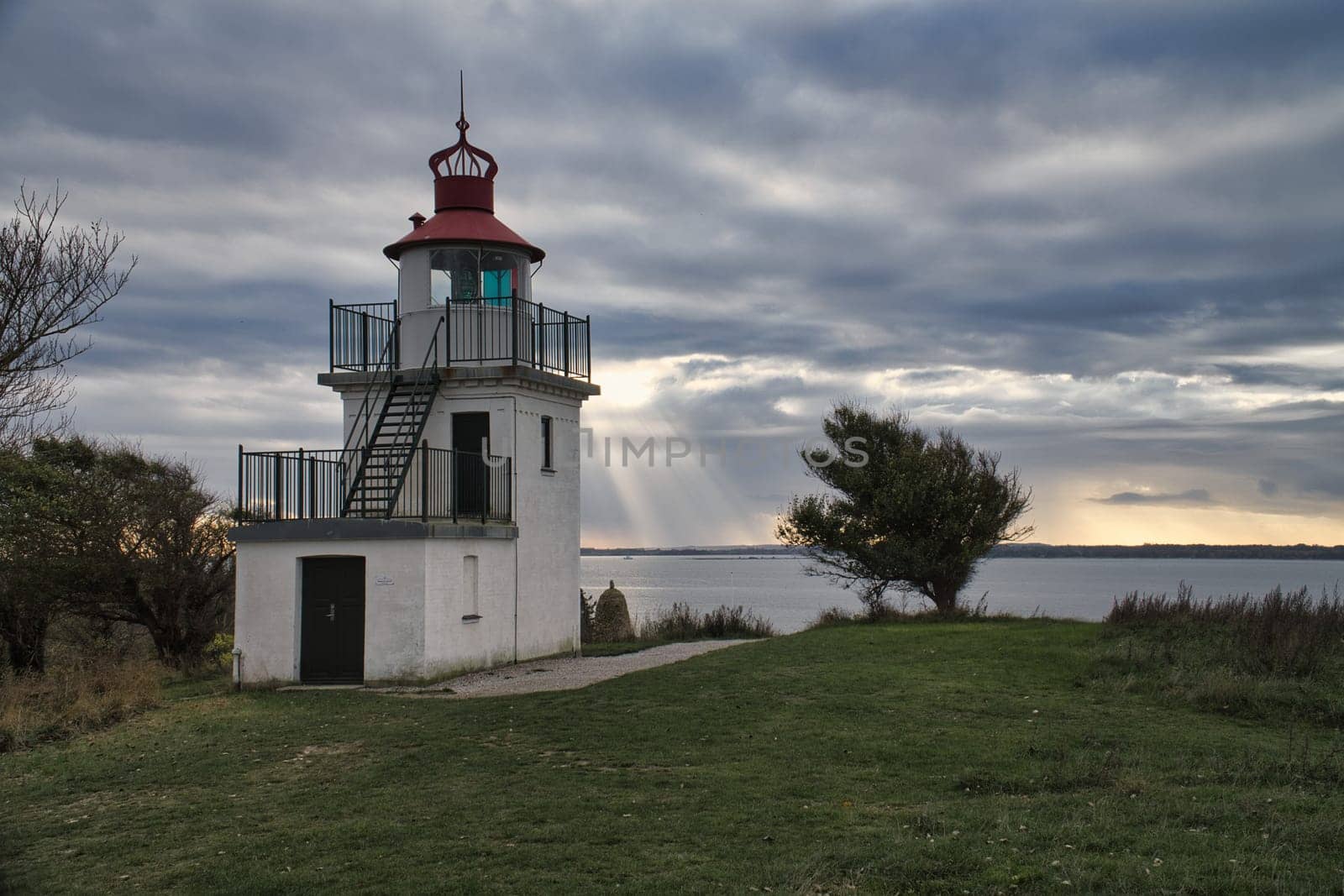 Lighthouse, Spodsbjerg Fyr in Huntsted on the coast of Denmark. Sun rays shining through the clouds. Meadow with trees. Landscape photo from sea