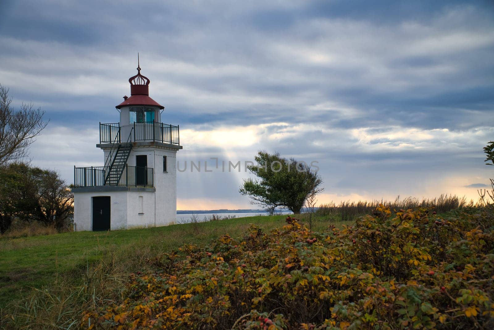 Lighthouse, Spodsbjerg Fyr in Huntsted on the coast of Denmark. Sun rays shining through the clouds. Meadow with trees. Landscape photo from sea