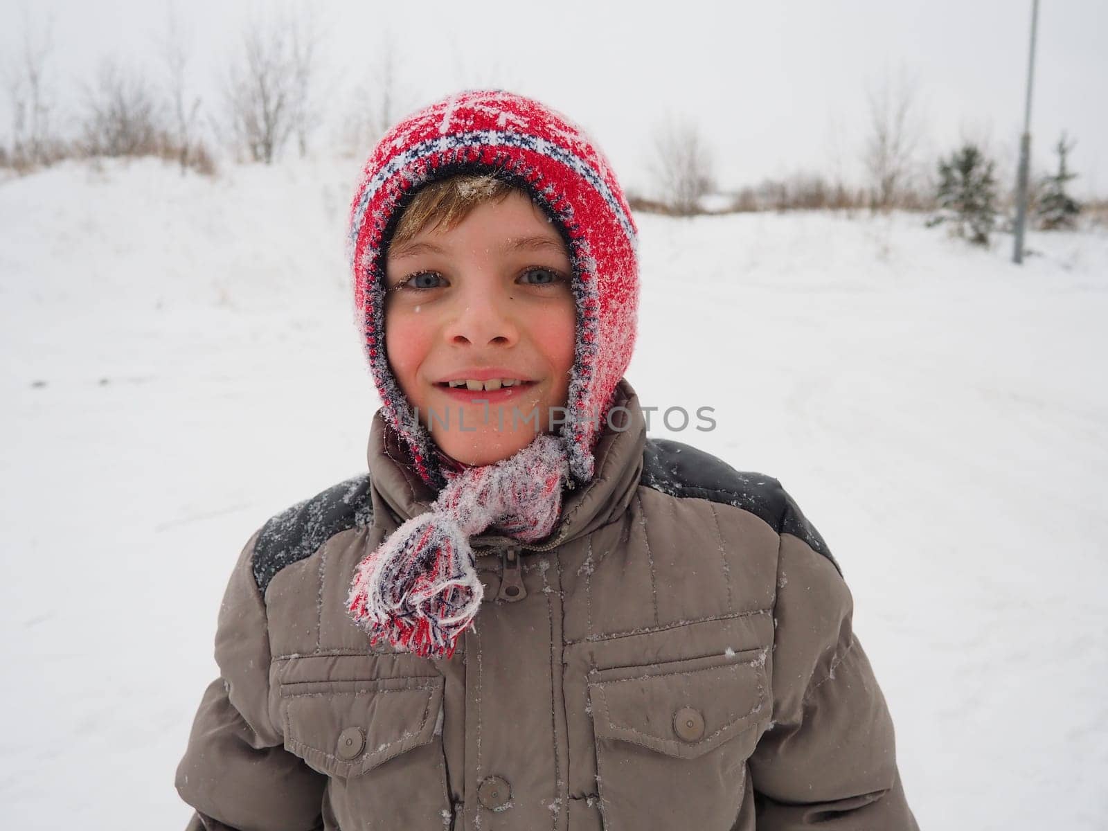 A cute boy 8 years old in a knitted red hat with braids and tassels. The child smiles cheerfully. Snowflakes on the face. The theme of winter holidays, frosty weather and travel in the north