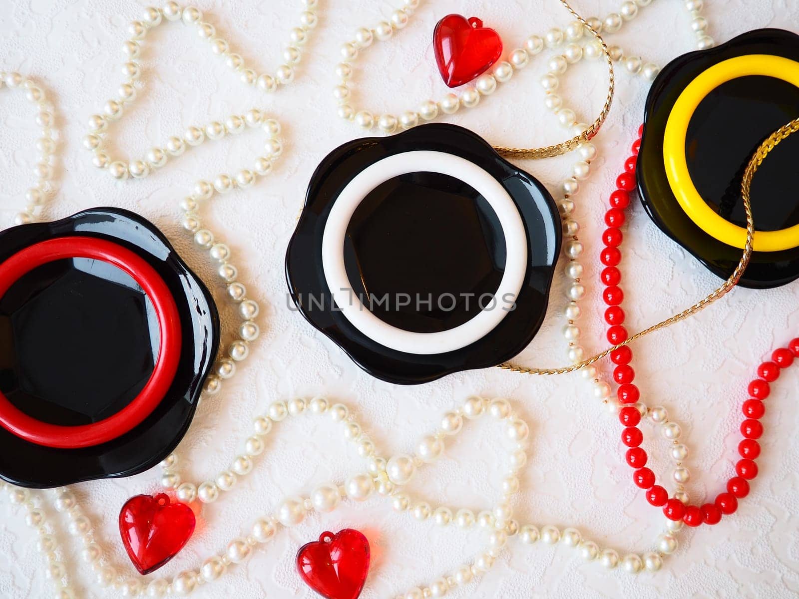 Flat lay. A beautiful composition of glass black dishes, plastic jewelry, bracelets, beads, red hearts on a white background. Women's baubles for February 14, Valentine's Day. Still life.