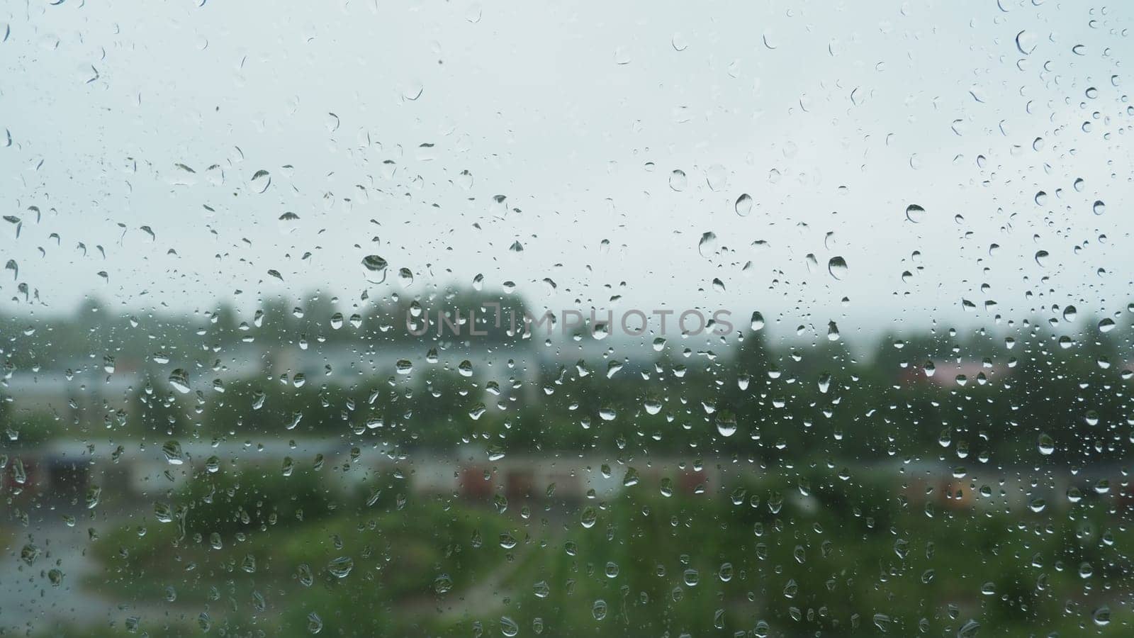 Raindrops on the glass window on a rainy day. Water drips from above. Blurred background. Street with burnings and trees outside the window. It's raining in the summer