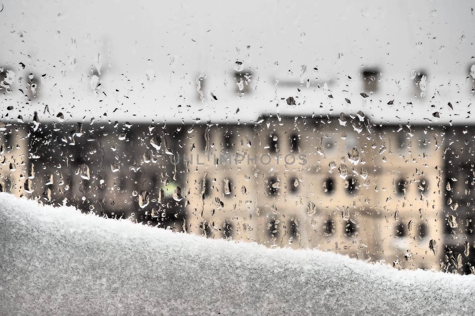 View from the window to the winter thaw. Melting snowdrift and drops on the glass. Multi-storey building with many windows. Christmas New Year postcard. Melancholy and tears or the arrival of spring